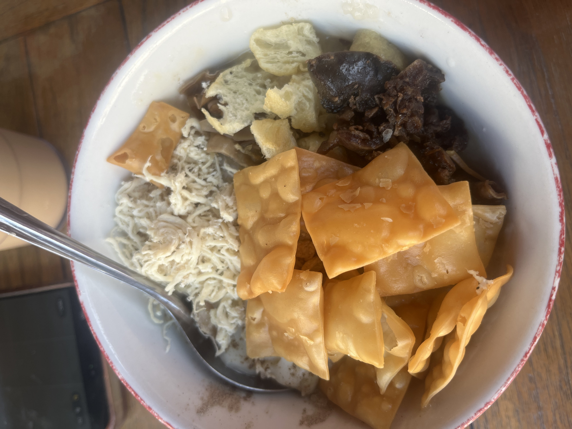 A close-up, top-down shot shows a bowl of noodles in Surabaya, Indonesia, with a spoon resting on top. The noodles appear to be a light color, possibly white or yellow, and are topped with shredded meat or egg, and crispy fried wonton wrappers. The bowl has a red and white striped rim and sits on a wooden surface, likely a table. The background is blurred but suggests an indoor setting with a smartphone and a beverage cup visible. The lighting is natural, coming from above, creating soft shadows.