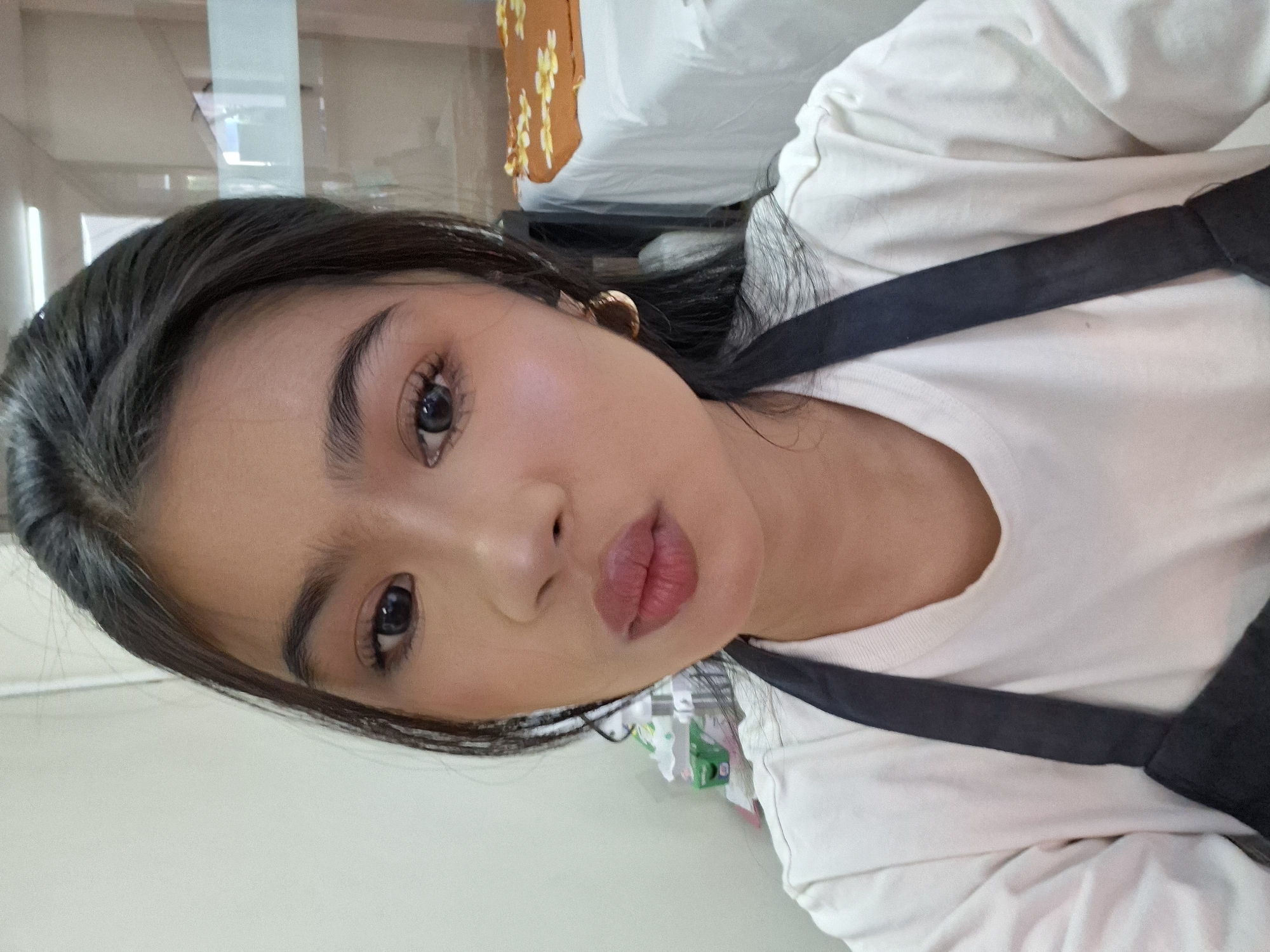 In a close-up, self-taken photograph, a young woman with dark hair pulled back from her face and a white t-shirt with a black apron is positioned in the foreground. She has on makeup, including eyeshadow, mascara, and lipstick, and is wearing a gold hoop earring in her right ear. Her lips are pursed. In the blurred background, shelves with boxes and a bed with a patterned blanket are visible, suggesting an indoor setting, possibly a salon or spa. The lighting appears to be artificial. The location is Kuta, Indonesia.