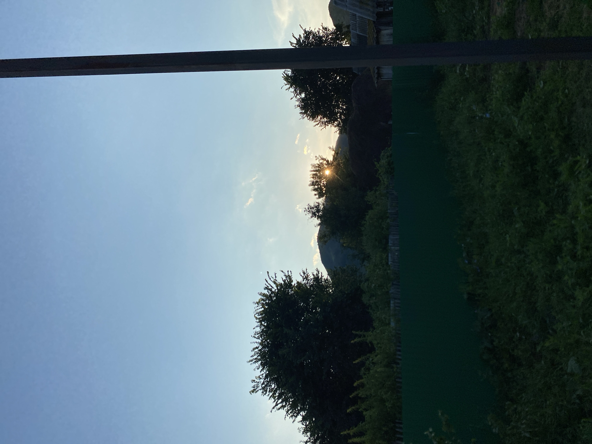 In Maloarslangulovo, Russia, a rural landscape unfolds under a clear sky at dusk. The horizon is dominated by the silhouettes of trees and a distant mountain range. The sun, partially obscured by the trees, casts a warm glow, creating rays of light that pierce through the foliage. In the foreground, a dark green fence runs horizontally across the frame, separating a grassy area from the more densely vegetated background. To the right, a vertical, dark pole stands prominently, its texture rough and weathered. The scene is tranquil, depicting a serene evening in the countryside.