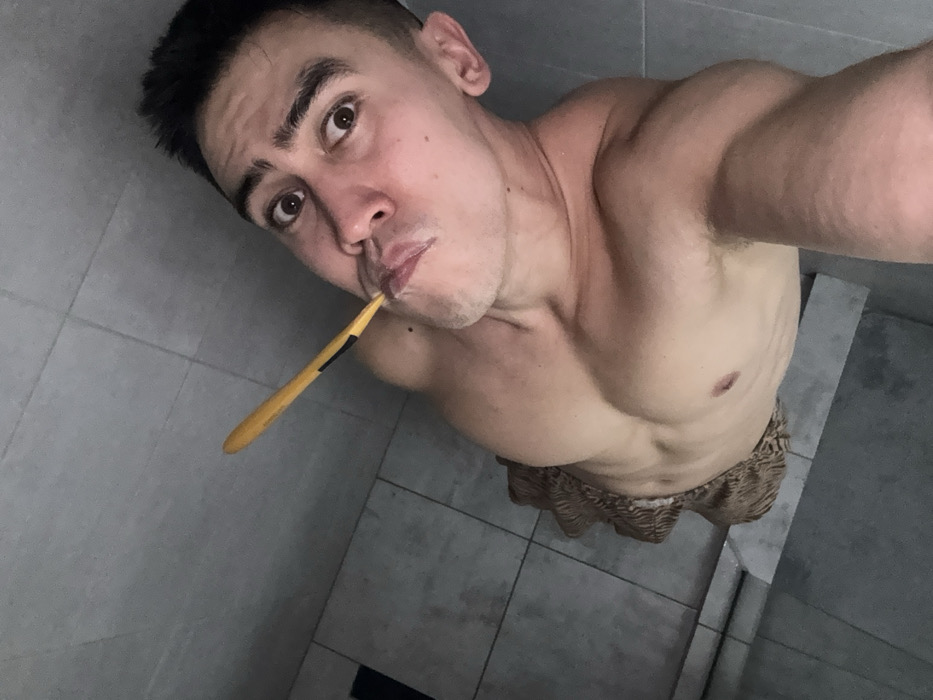A man takes a selfie in a bathroom, shirtless and holding a toothbrush in his mouth. The bathroom has gray tiled walls and floors. He's looking up at the camera. His facial expression appears neutral. The toothbrush has a yellow handle. He is wearing patterned shorts. The lighting in the bathroom is somewhat dim.