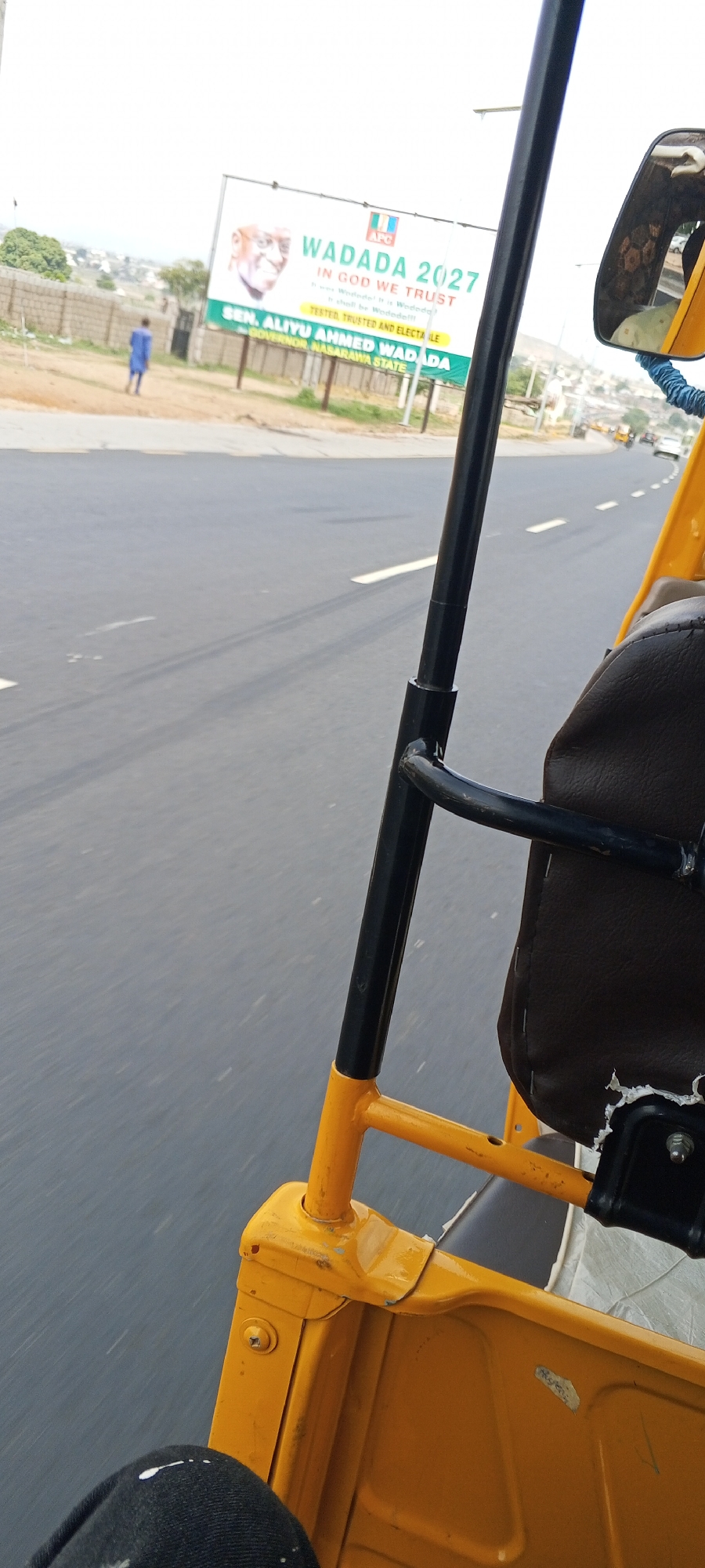The image shows a view from inside a yellow auto-rickshaw, commonly known as a "Keke" or "Tricycle", moving along an asphalt road. The road has white lane markings. On the roadside, there is a billboard promoting "WADADA 2027" with the text "IN GOD WE TRUST", and "SEN. ALIYU AHMED WADADA GOVERNOR NASARAWA STATE". A person is walking near the billboard. The location is Uke, Nigeria.