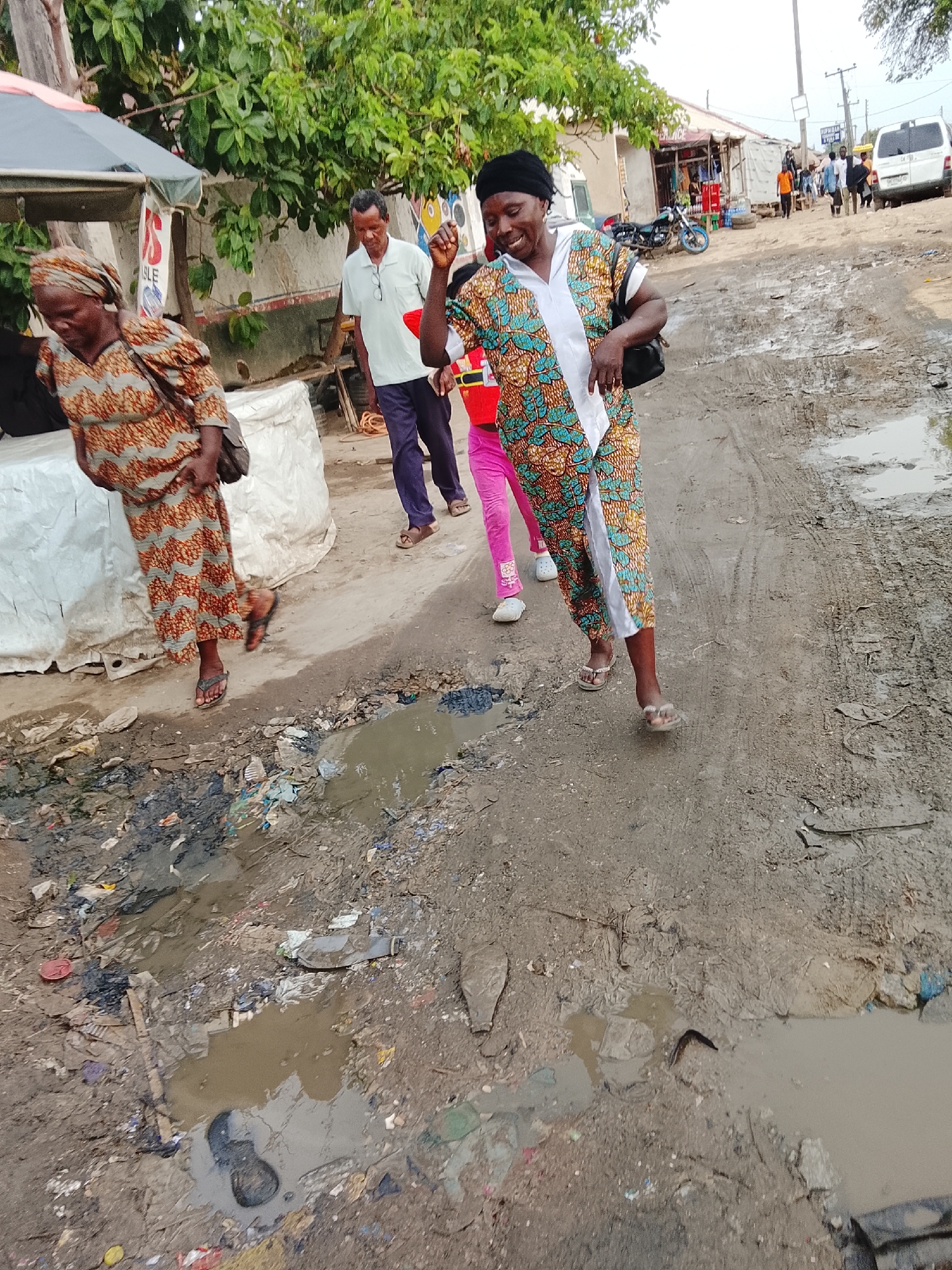The image depicts a muddy and debris-strewn street scene. In the foreground, there are puddles of water mixed with trash and discarded items. Four people are walking on the street, presumably attempting to avoid the worst of the mud and debris. The woman in the foreground wears a colorful patterned dress with a white front panel, and a black head covering. An older woman walks ahead of her in a brown patterned dress. A man in a white shirt and purple pants follows behind them with a child in pink pants. In the background, there is a building with signs, a parked motorcycle, other people, and a white van. Based on the visible signage and architectural style, the location is in Abuja, Nigeria.