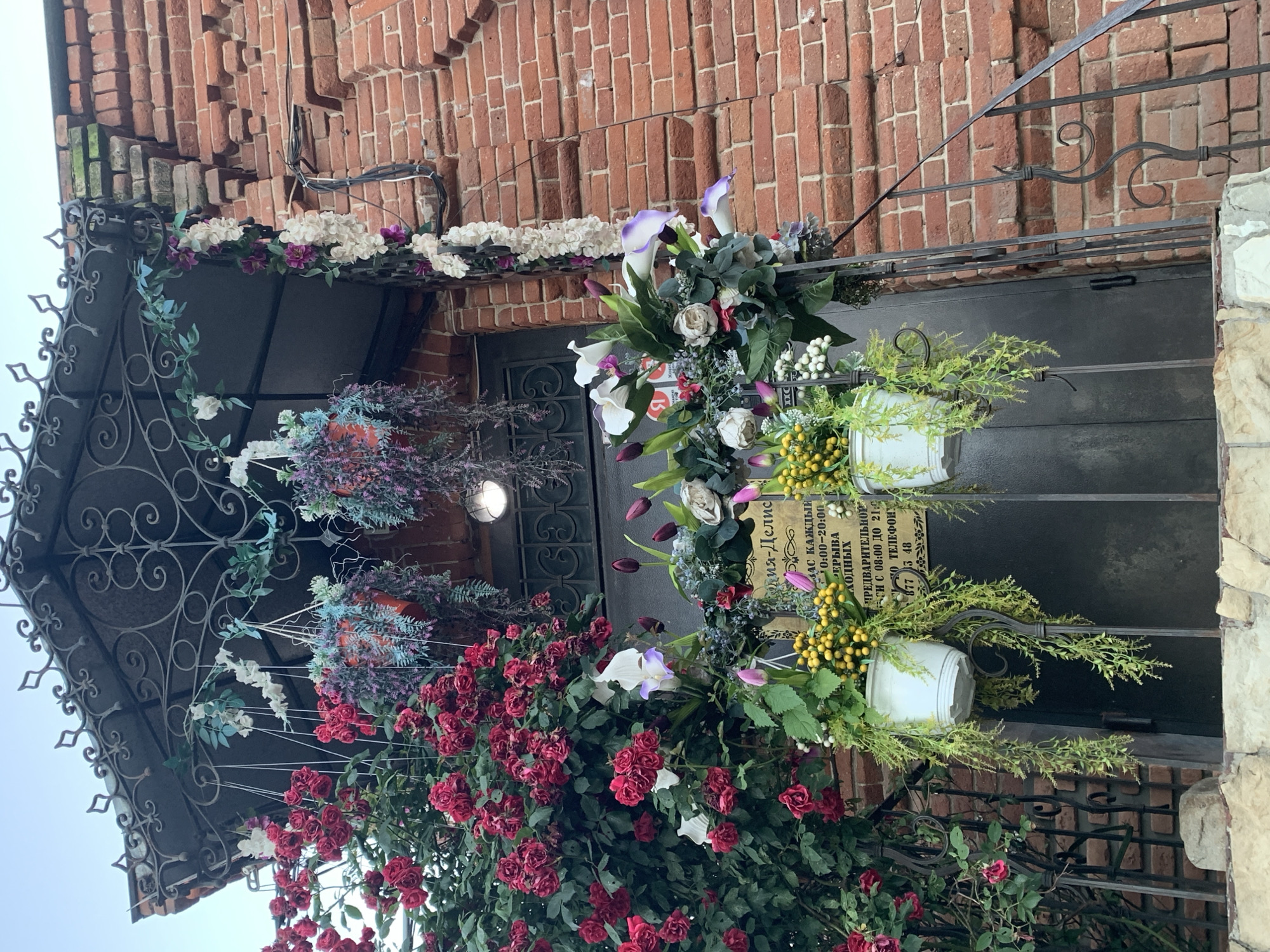 An exterior medium shot captures a brick building adorned with abundant floral decorations. Red roses cascade down the left side of the image, while hanging baskets filled with artificial greenery and flowers are suspended from an ornate metal overhang. Below the overhang, a door is flanked by two white planters containing more artificial flowers. A sign with Russian text is visible on the door. A black metal railing extends down the right side of the frame, partially obscured by the vegetation and flowers. The brickwork of the building appears aged and weathered. The sky above is a pale, overcast gray. There are no people or animals visible in the scene.