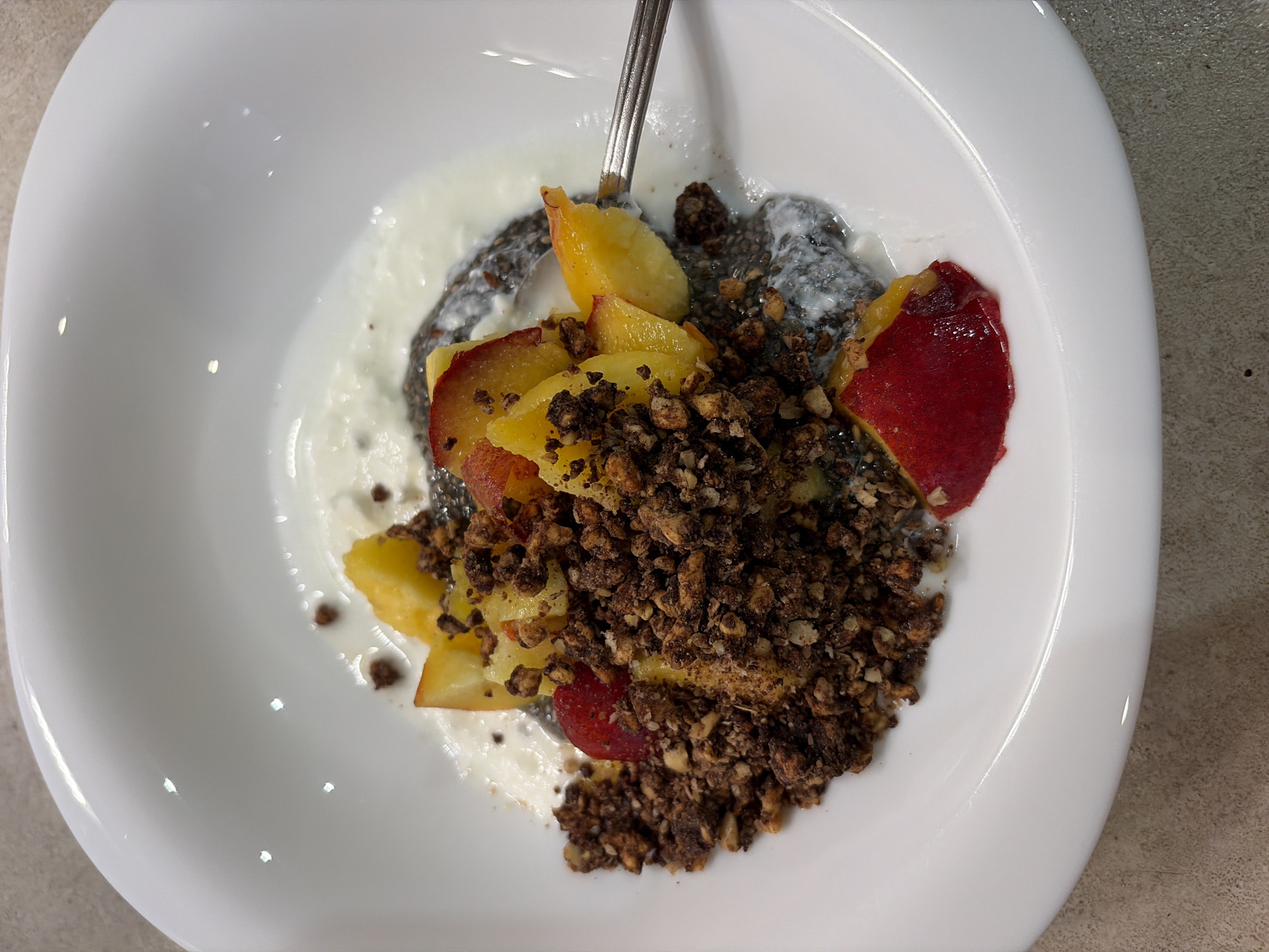 A close-up, overhead shot depicts a white, oblong ceramic bowl filled with a healthy breakfast or dessert, set against a light-colored countertop. The bowl contains a creamy white base, likely yogurt, topped with a dark, speckled layer resembling chia seed pudding. Freshly sliced fruit, appearing to be peaches or nectarines, with their vibrant yellow-orange flesh and reddish skin, are arranged over the creamy layers. A generous portion of dark brown, coarse granola or crumble, possibly chocolate-flavored or containing nuts, covers the center of the dish. A silver metal spoon is partially submerged into the food, suggesting it is either being prepared or consumed. The scene is brightly lit, highlighting the varied textures and colors of the ingredients. The overall presentation is appealing, indicative of a fresh and wholesome meal. The image was captured in Burgas, Bulgaria.