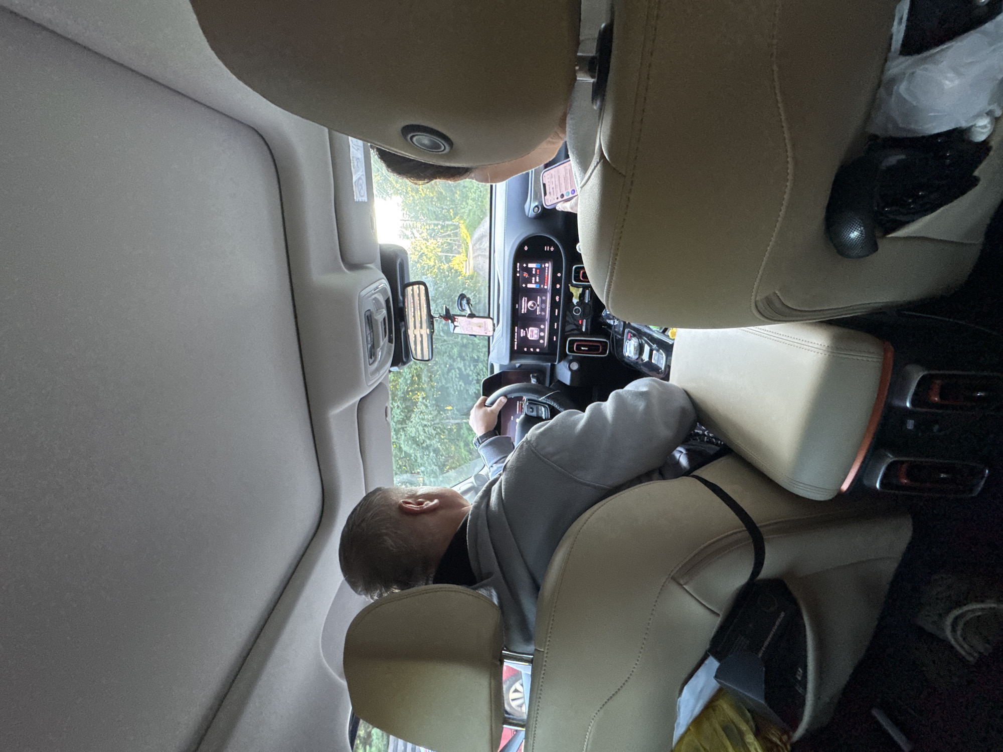 The image depicts the interior of a car, likely in Zelenaya Roshcha, Russia, as viewed from the back seat. A man with short gray hair is driving, wearing a gray hooded sweatshirt and a watch on his left wrist. He is holding the steering wheel with both hands. In the passenger seat, a person is visible from the back, also wearing a light-colored top, and holding a smartphone.
The car's interior features tan leather seats, a large infotainment screen on the dashboard displaying various icons and information, and a rearview mirror with a hanging air freshener and smartphone mount. The dashboard also has climate control vents and buttons. Sunlight streams in through the windshield and a sunroof, illuminating the interior. Outside the car, trees and foliage suggest a natural or suburban setting, possibly a road surrounded by greenery. The overall atmosphere is casual, with personal items such as a black box and plastic bags visible in the rear of the car.
