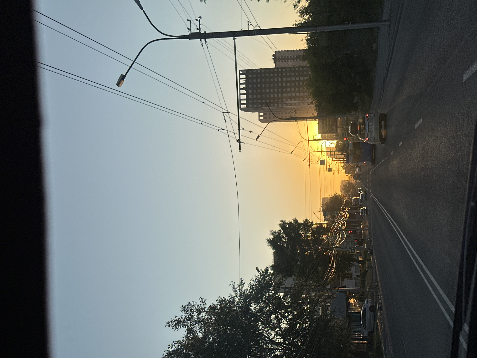 The image depicts a street scene in Novosibirsk, Russia, during what appears to be sunset or sunrise. The perspective is from inside a vehicle, looking forward. A road stretches into the distance, marked with double white lines. On the left side of the road, there are trees and buildings, including a white car parked near a building. Traffic lights are visible, and several cars are driving on the road. On the right side of the road, there are more trees and utility poles with overhead wires, along with tall buildings in the background. The sky is clear, transitioning from a light blue at the top to a warm orange-yellow hue near the horizon where the sun is setting or rising, casting a glow over the scene.
