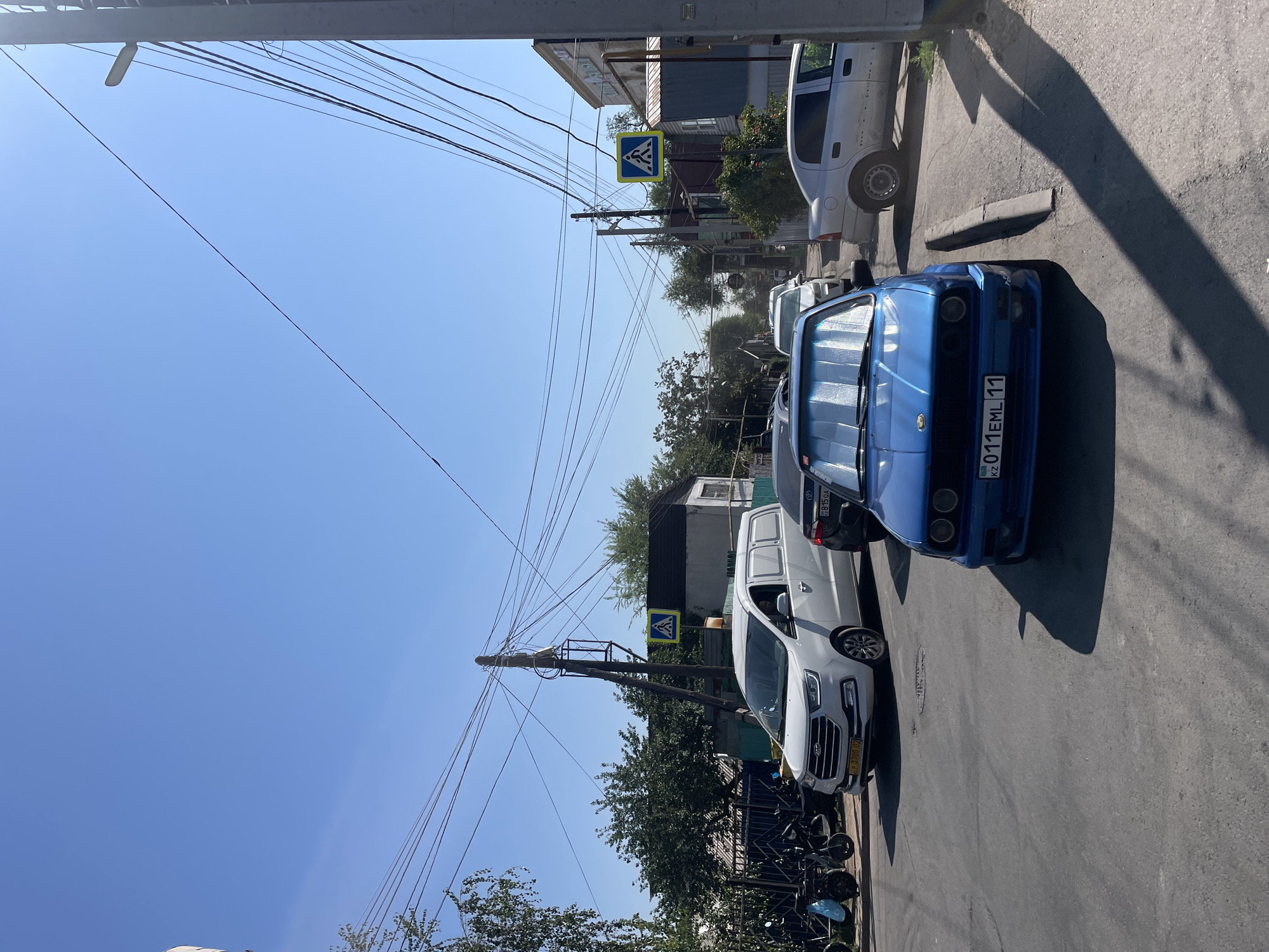 The image depicts a street scene in Almaty, Kazakhstan, on a sunny day. The foreground is dominated by a blue BMW car, its front visible, with a license plate reading "KZ 011EML 11". Behind it, a white van with the license plate "P 3708 02" is stopped, and a black car is partially visible. Further down the street, a silver car is parked. The street is paved, with a curb on the right side. Overhead, a dense network of electrical wires stretches across the scene, supported by utility poles. There are two pedestrian crossing signs visible on poles. Trees and buildings line the sides of the street, including a building with some signage in Cyrillic. On the left side of the street, several parked motorcycles or scooters are visible. The sky is clear and blue. The overall impression is that of a typical urban street with moderate traffic.