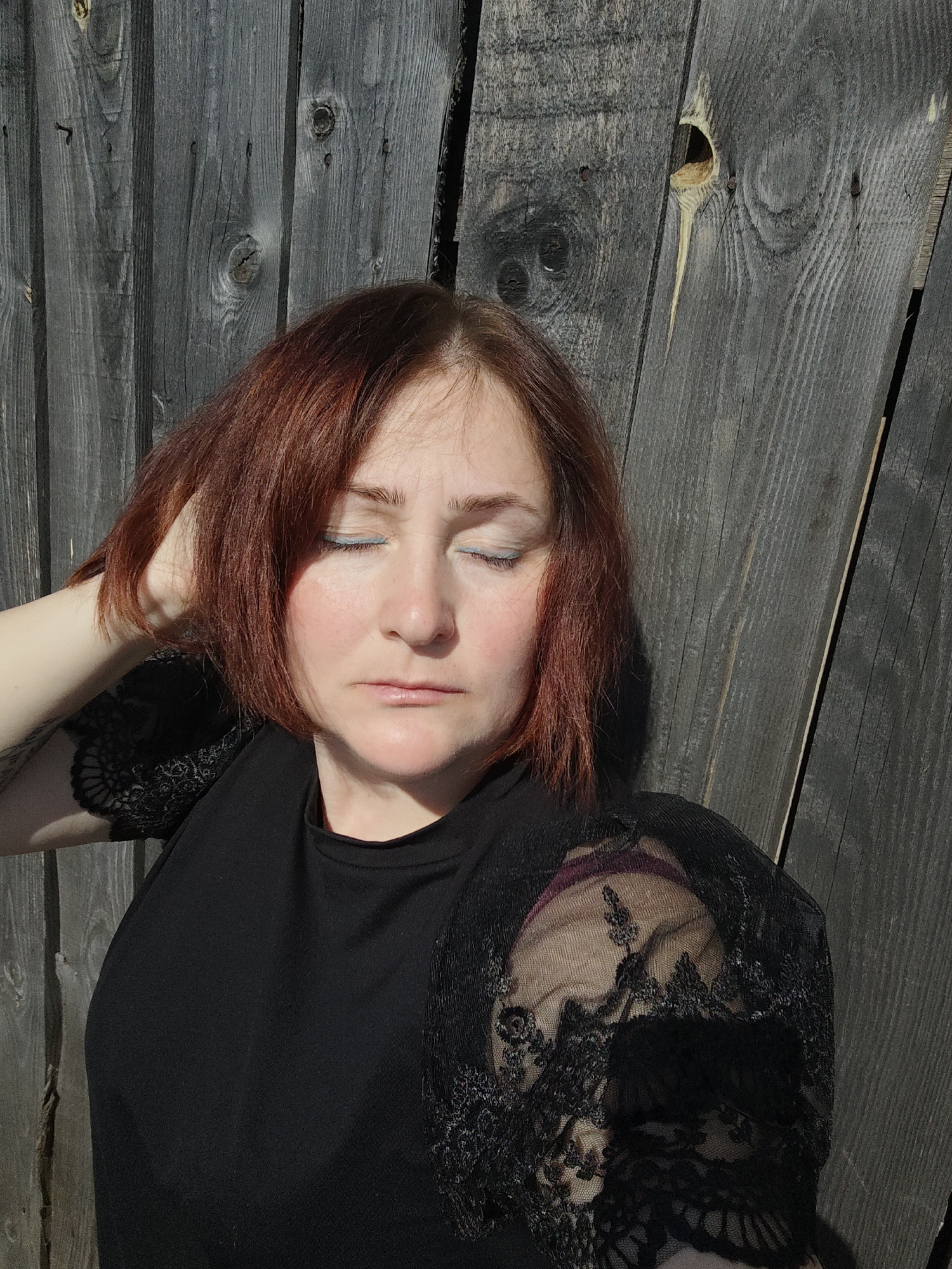 A woman with red-brown hair and pale skin stands against a weathered wooden fence. She wears a black top with lace sleeves. Her eyes are closed, and her expression is serene. The lighting suggests a sunny day, casting shadows on the fence and her face. The background is the gray wood of the fence. The city of Фоки, Russia is not discernible from this image.