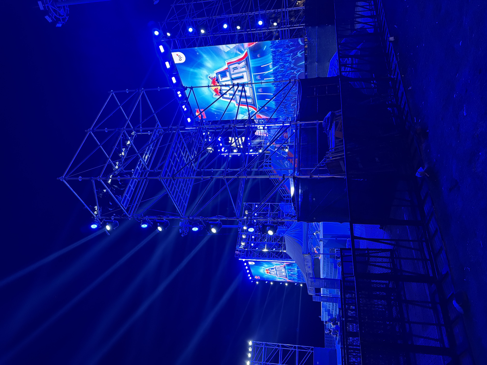 A night scene depicts a large outdoor stage structure illuminated by bright blue lights and spotlights. A massive LED screen displays a vibrant graphic with the text "RED BULL GC TOUR" and the Red Bull logo. Scaffolding surrounds the stage, and additional lighting rigs are visible. In the foreground, a black metal barrier separates the viewer from the stage area. The ground is dark and appears to be dirt or gravel, with some debris scattered around. The overall impression is that of a concert or event setup at night. The location of the image is stated to be Nha Trang Ward, Vietnam.