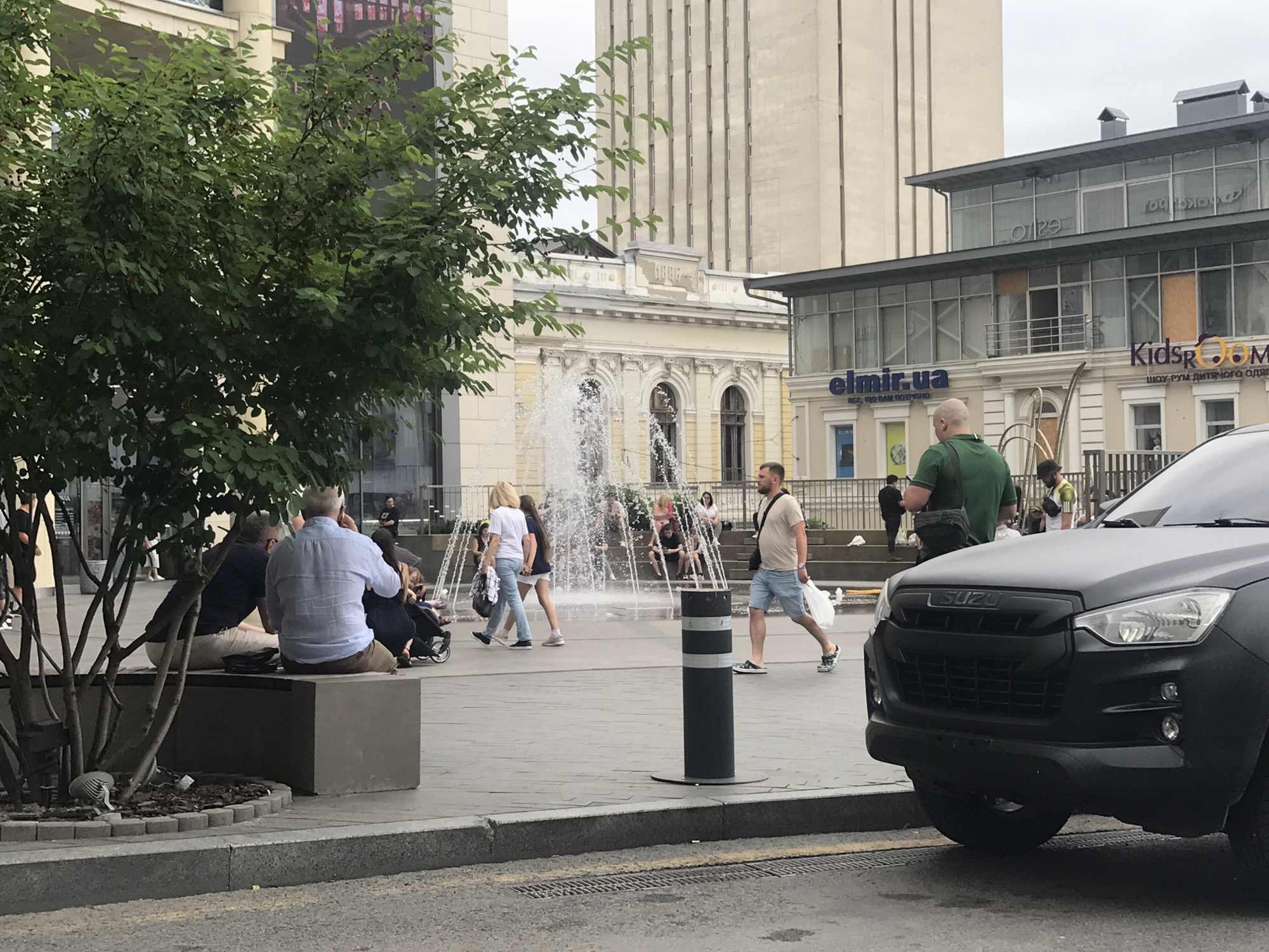 The image depicts an outdoor urban scene in Kharkiv, Ukraine, likely in the vicinity of the "El Miron" shopping center. In the foreground, a matte black SUV, identified as an Isuzu, is parked. To the left, a tree with green foliage and small red berries provides shade over a concrete bench where two men are seated. One man wears a light blue shirt and light brown pants, while the other is in a dark blue shirt and light-colored pants. Further back, a fountain is active, with water spraying upwards. Several people are visible near the fountain, including a woman in a white top and blue jeans walking to the left, and a man in a light brown t-shirt and blue shorts walking to the right, carrying a white bag. Behind them, there are buildings with modern architectural elements, including large glass windows, and some older facades with decorative elements. The ground is paved with grey tiles. The overall atmosphere appears to be daytime, with an overcast sky.