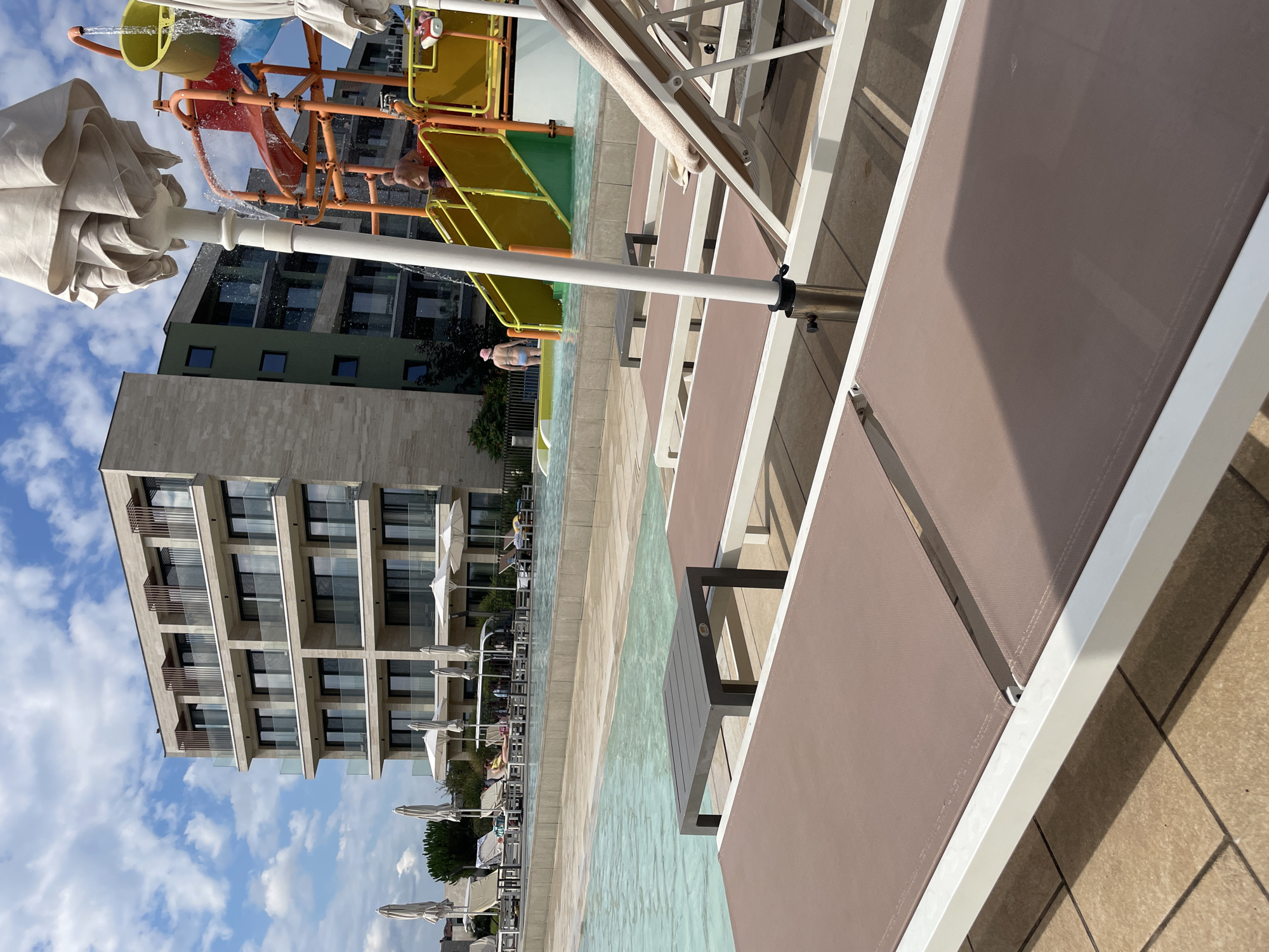 This is a photograph taken at a resort in Obzor, Bulgaria, depicting a poolside scene on a sunny day. In the foreground, several empty, modern lounge chairs with brown fabric are arranged on a tiled patio. A small, dark gray rectangular table sits between two of the lounge chairs. Beyond the lounge chairs, a shallow, clear blue swimming pool is visible, with a tiled edge.
In the middle ground, a multi-story hotel building with a contemporary architectural style, featuring balconies and beige stone cladding, stands against a backdrop of a partly cloudy blue sky. To the right, a colorful water slide structure with yellow and orange elements is present, with a person in a pink hat and blue swimwear standing near its base and another person, shirtless, on the stairs of the slide. Several white umbrellas are also scattered around the poolside area. The environment suggests a recreational setting, likely a hotel or resort complex.
