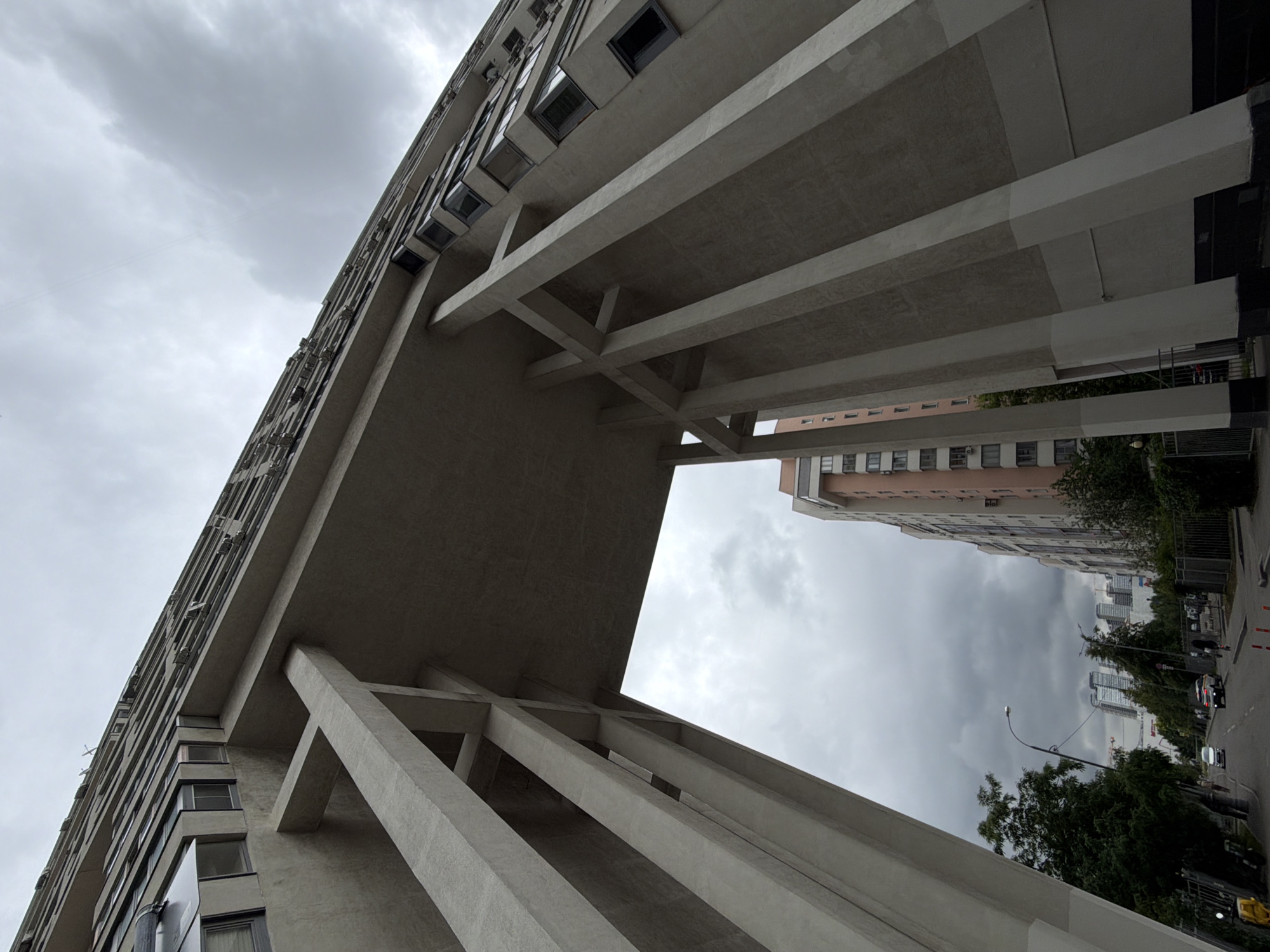 This low-angle, wide-shot photograph captures the Brutalist architecture of a residential building under a cloudy, overcast sky. The building is made of concrete, with a series of cantilevered sections supported by large, angular columns. Rows of windows are visible along the facade, many with their vents open.
In the background, another building with a pinkish facade can be seen, along with glimpses of trees and a street with cars. The overall scene is somewhat stark and geometric, emphasizing the concrete structures. The weather appears to be gloomy, with dark clouds dominating the sky. There is no visible text in the image. The setting appears to be an urban environment, likely a residential area in Khimki, Russia, based on the provided location context. No people are clearly visible, and no specific activity or event is taking place. The time of day is difficult to ascertain precisely due to the overcast conditions, but it suggests daytime. The mood is somewhat somber, reflecting the weather and the monolithic architecture.
