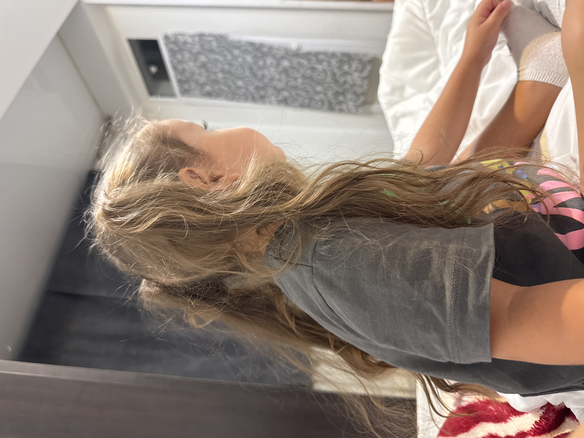 The image shows a young girl from the side, her face turned away from the camera. She has long, blonde hair styled in two pigtails. She is wearing a dark grey t-shirt. The girl is sitting on a white bed with white bedding. To the left, a blanket with a red and white striped pattern is visible. In the background, a dark curtain hangs to the left, and a doorway with a patterned curtain is visible on the right. The lighting suggests an indoor setting, possibly a bedroom. There are no discernible landmarks or environmental cues that would allow for a precise location within Chișinău, Moldova.