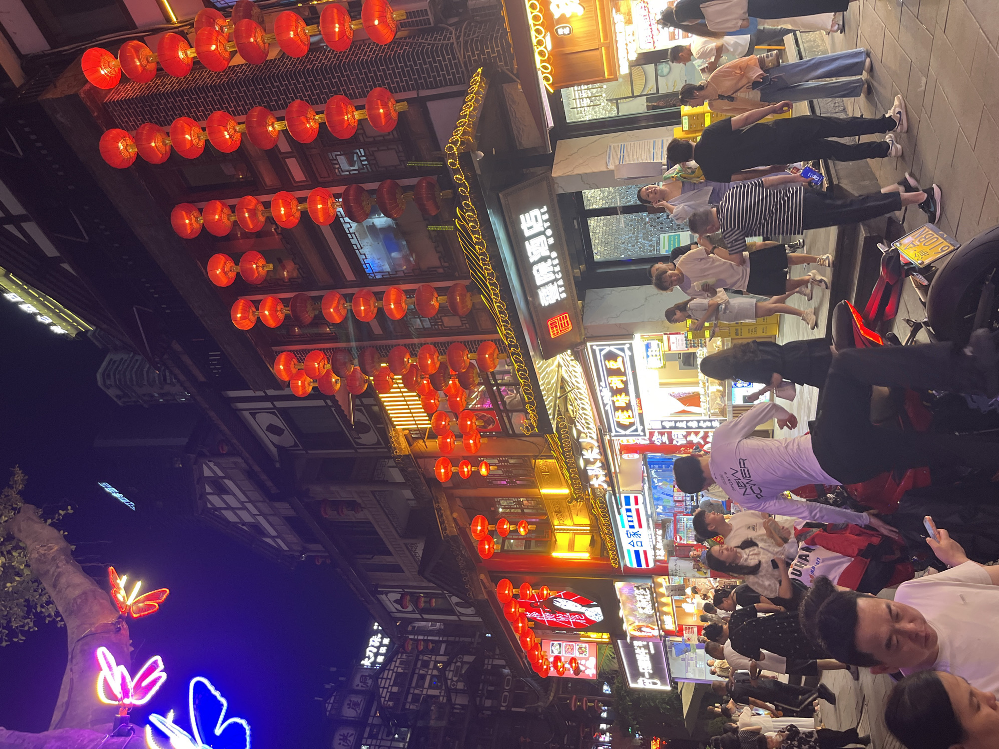A nighttime scene on a bustling street in the Yuzhong District of China shows a multi-story building adorned with traditional Chinese architecture, illuminated by numerous red lanterns. Neon signs and various storefronts are visible, some with Chinese characters and English translations. In the foreground, a group of people are gathered on a tiled sidewalk. One person, wearing a white long-sleeved shirt with "SPORT'S NEW POWER" printed on it, is interacting with a red motorcycle. Another individual in the foreground is looking at a smartphone. Further back, several other people are walking, conversing, and looking at the shops. The overall atmosphere suggests a vibrant commercial area during the evening.