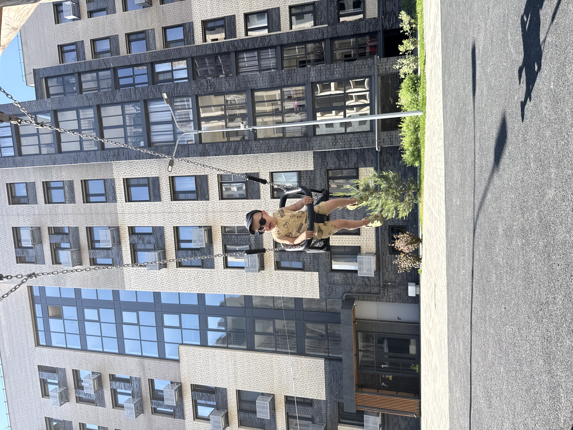 In Rusanovo, Russia, a young boy wearing a black baseball cap, sunglasses, and a light-colored patterned jumpsuit is swinging on a black swing set in front of a modern, multi-story apartment building. The building features a facade of light beige and dark grey bricks, with numerous windows. The boy is mid-swing, with his legs extended, and appears to be looking forward. The swing set is supported by thick metal chains. In the foreground, a paved area with a textured asphalt surface is visible, with a shadow of a street lamp cast across it. Green bushes and a small evergreen tree are planted to the right of the building's entrance, which is marked by a dark canopy and a sign with the number "1." The sky above is a clear blue.