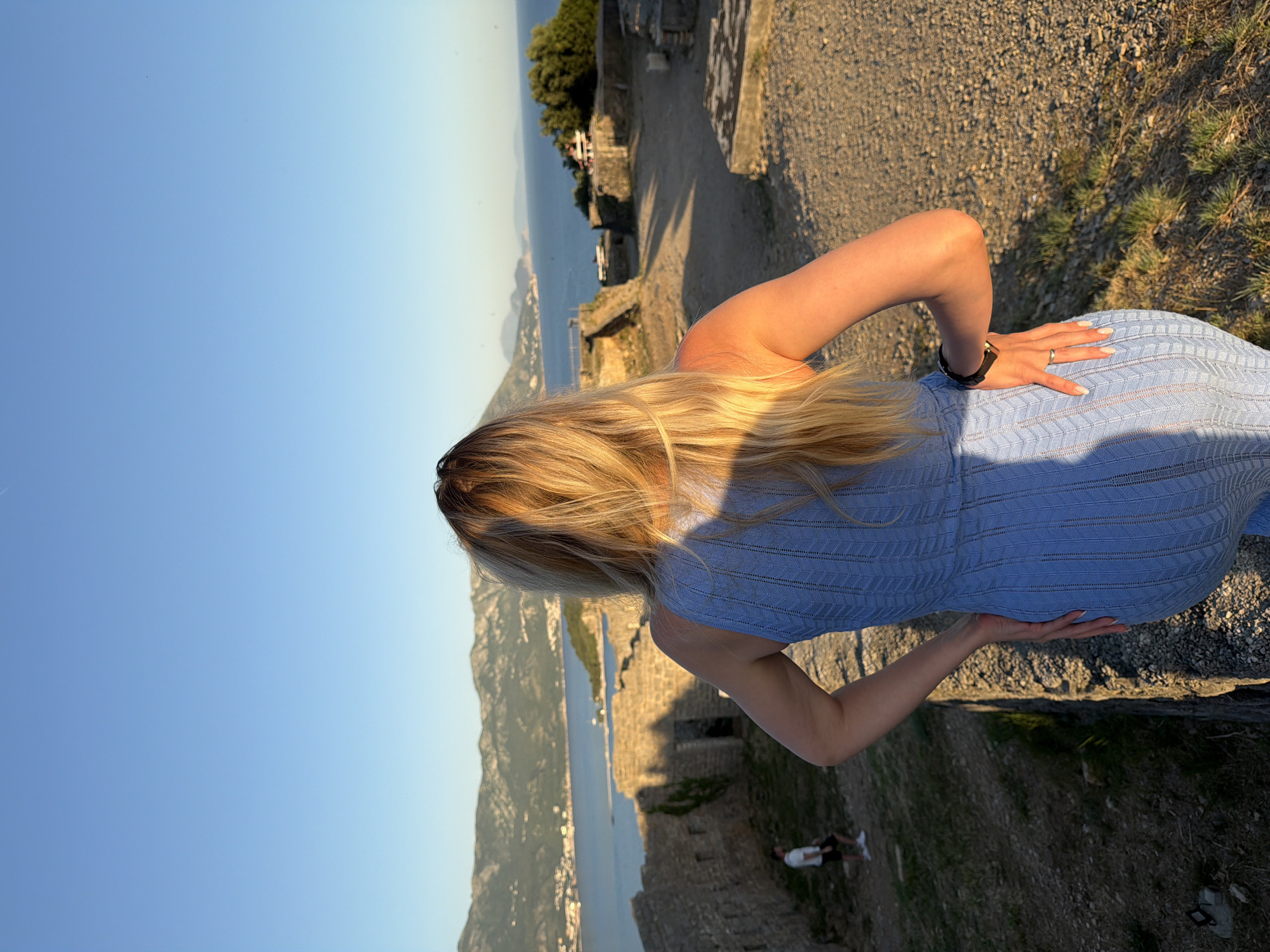 This image captures a woman from behind, standing on elevated ground within ancient stone ruins, looking out over the coastal landscape of Budva, Montenegro, during the golden hour. The woman has long, sun-kissed blonde hair, and wears a light blue, sleeveless, form-fitting dress with a patterned knit. Her left arm is bent with her hand on her hip, revealing a dark-strapped watch on her wrist and a ring on her left ring finger; her right hand is also on her hip. In the mid-ground, further stone fortifications and structures are visible, with a man in a white t-shirt and dark shorts walking amongst them on the left. The background features a expansive bay of the Adriatic Sea, bordered by mountains and the clustered buildings of the Budva Riviera. The sky is clear blue, and the low-angle sunlight casts warm, golden hues and long shadows across the scene.