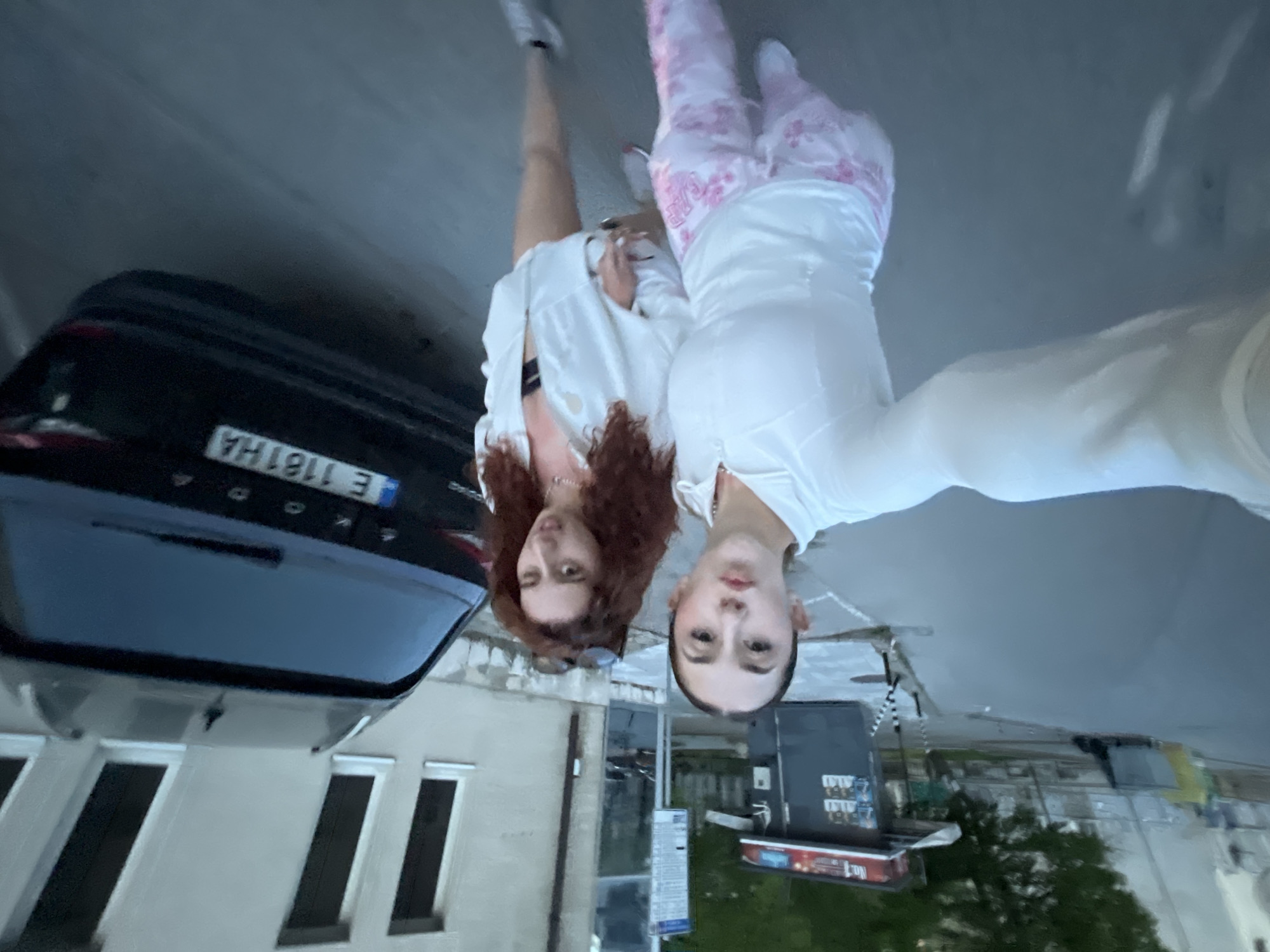 Two females take a selfie in Blagoevgrad, Bulgaria. One female is on the left, wearing a white top and shorts, with sneakers, and red curly hair. The other female is on the right, wearing a white long-sleeved top and pink floral patterned pants. The license plate on the car reads "E 1181 HA." The background includes buildings and greenery. The photograph is taken with an upside-down orientation.