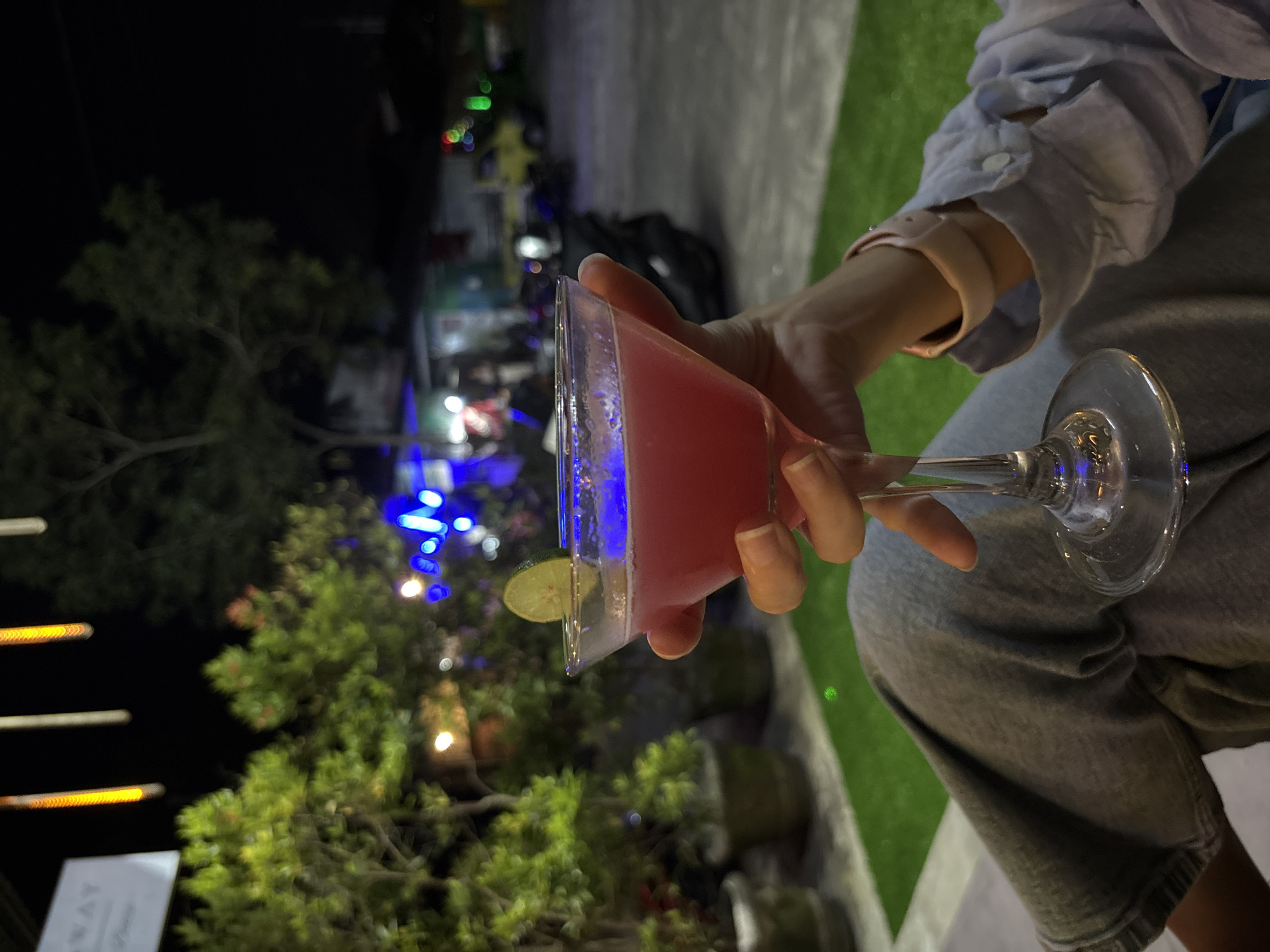 A person holds a triangular glass filled with a pink liquid, likely a cocktail, garnished with a slice of lime. The person wears a light blue, button-down shirt, a light pink watch, and gray pants. The background is blurry but suggests an outdoor setting at night, with green foliage, string lights, and other lighting fixtures visible. The location could be in Kuta Selatan, Indonesia, based on the tropical plants and overall ambiance.