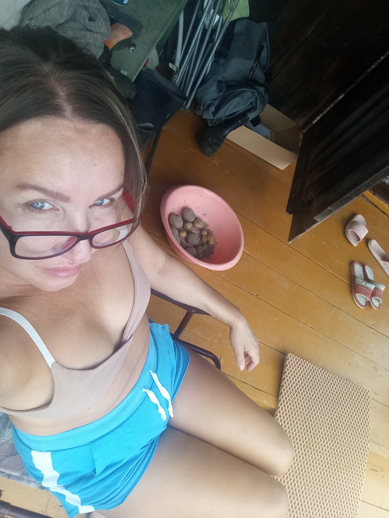 A woman with blonde hair and blue eyes, wearing glasses, a beige bra, and blue shorts, sits on a chair indoors. She is looking at the camera. In the background, there are various objects scattered around, including a pink bowl with stones, a pair of pink flip-flops, and a pair of striped slippers. The floor is made of wooden planks. The scene appears to be in a domestic setting.