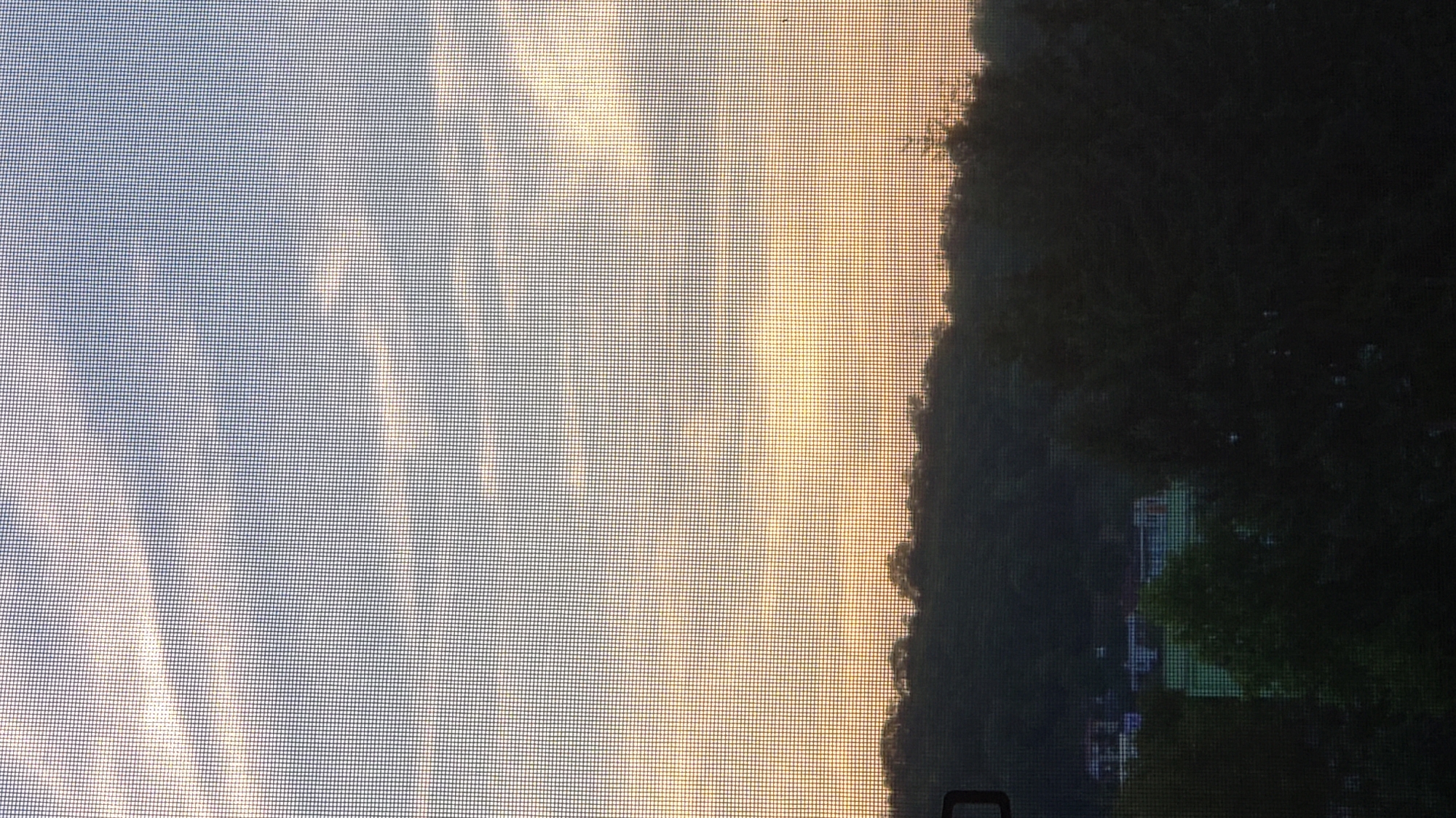 The image shows a sunset viewed through a window screen, with a silhouetted tree line and some buildings in the distance. The sky is a mix of blue, white, and orange, suggesting clouds are present and illuminated by the setting sun. The lower portion of the image is dominated by the dark silhouettes of trees and houses, indicating it is likely late evening. The scene is viewed from an elevated position looking out over a landscape that appears to be on the outskirts of a city. The presence of houses and what looks like a sports field or recreational area suggests a semi-urban or suburban environment. Without specific landmarks visible, pinpointing the exact location within Prešov, Slovakia is not possible, but the overall scene is consistent with a view from a residential area overlooking a wooded or hilly expanse.