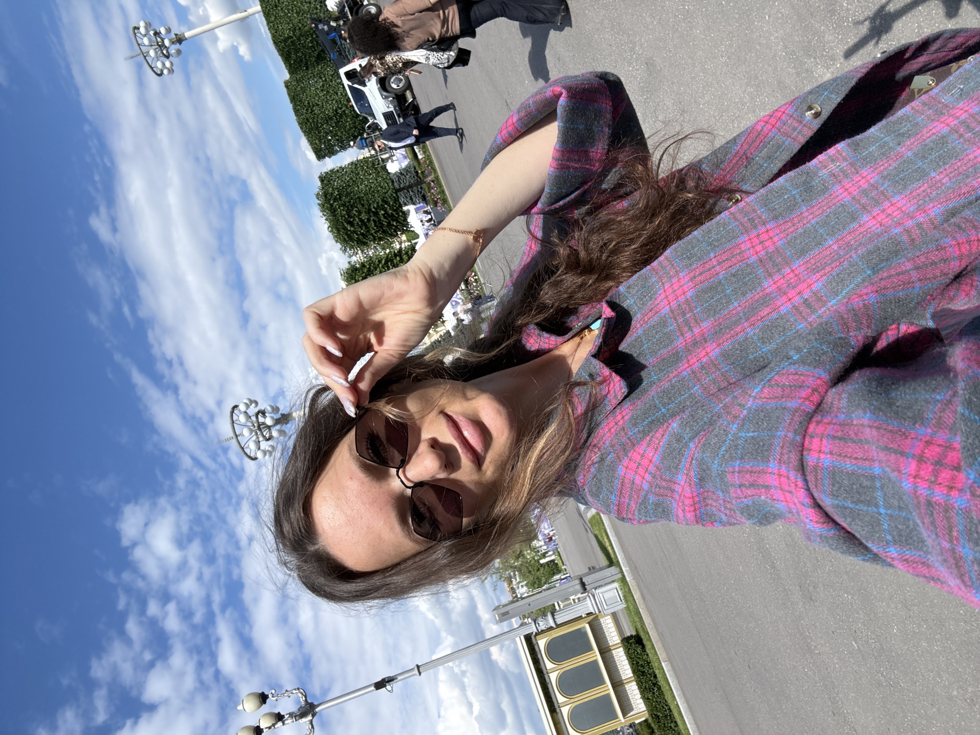 A woman is taking a selfie in an outdoor urban environment, possibly at an event or exhibition. She is wearing a plaid shirt with pink and grey colors and sunglasses, and she is adjusting them with her right hand. In the background, there are people walking, trees, a white truck, and lampposts. The sky is blue with white clouds. The architectural style of the building on the left, with its arched windows, and the presence of decorative lampposts are reminiscent of Soviet-era architecture often found in parks and exhibition grounds in Moscow, Russia, such as VDNKh.