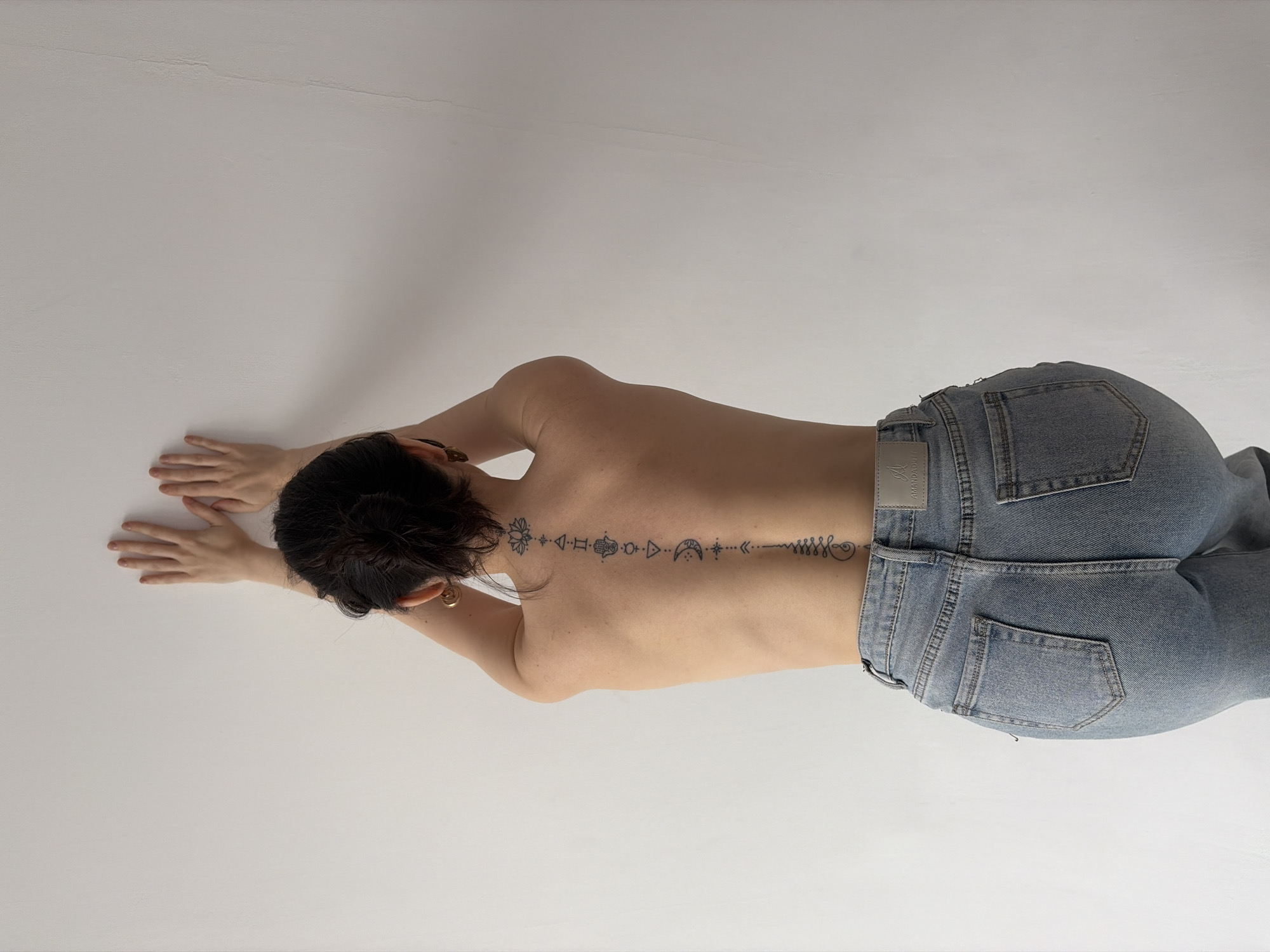 A woman wearing blue jeans lies prone on a white surface. Her upper back is bare and faces the camera. A tattoo extends down her spine. Her hands are outstretched in front of her.
The scene is indoors, and the location, Chaikovsky, Russia, is extraneous.