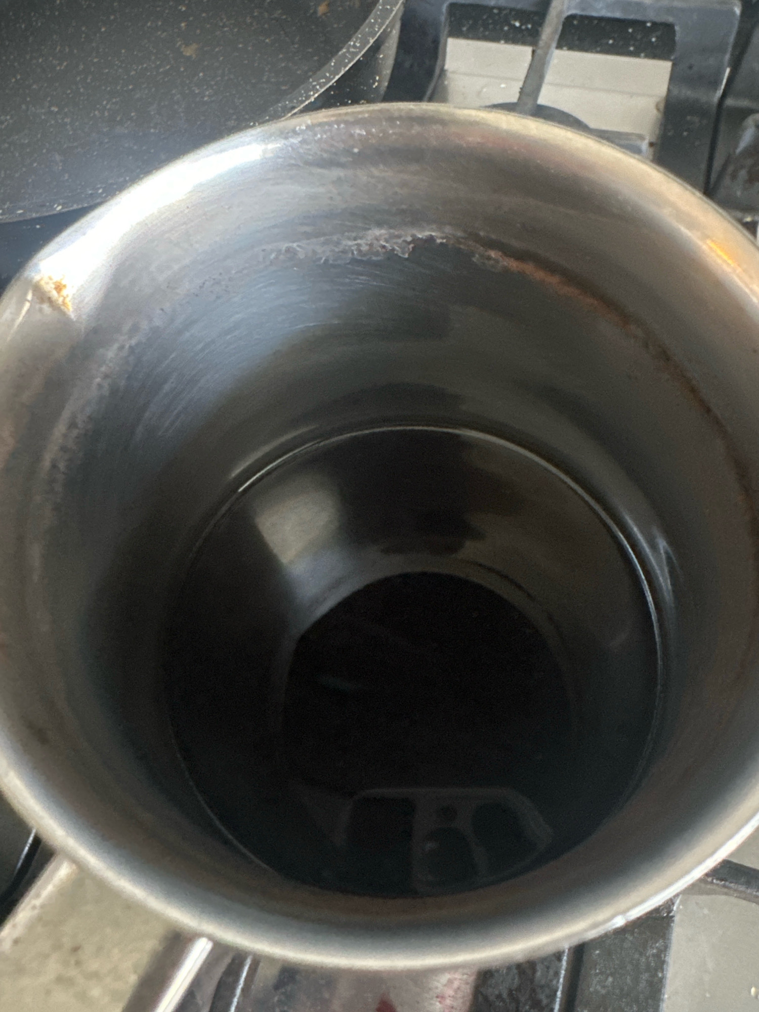 An extreme high-angle close-up shot shows the interior of a stainless steel pot containing a dark liquid. The pot has a slightly tarnished and scratched surface, with some brown discoloration visible along the rim and upper interior. The liquid reflects a distorted image of a cell phone camera. The pot appears to be sitting on a stovetop with a metallic grate. The scene is indoors, likely a kitchen. The image does not contain any information that can be used to determine its location in Nevinnomyssk, Russia.