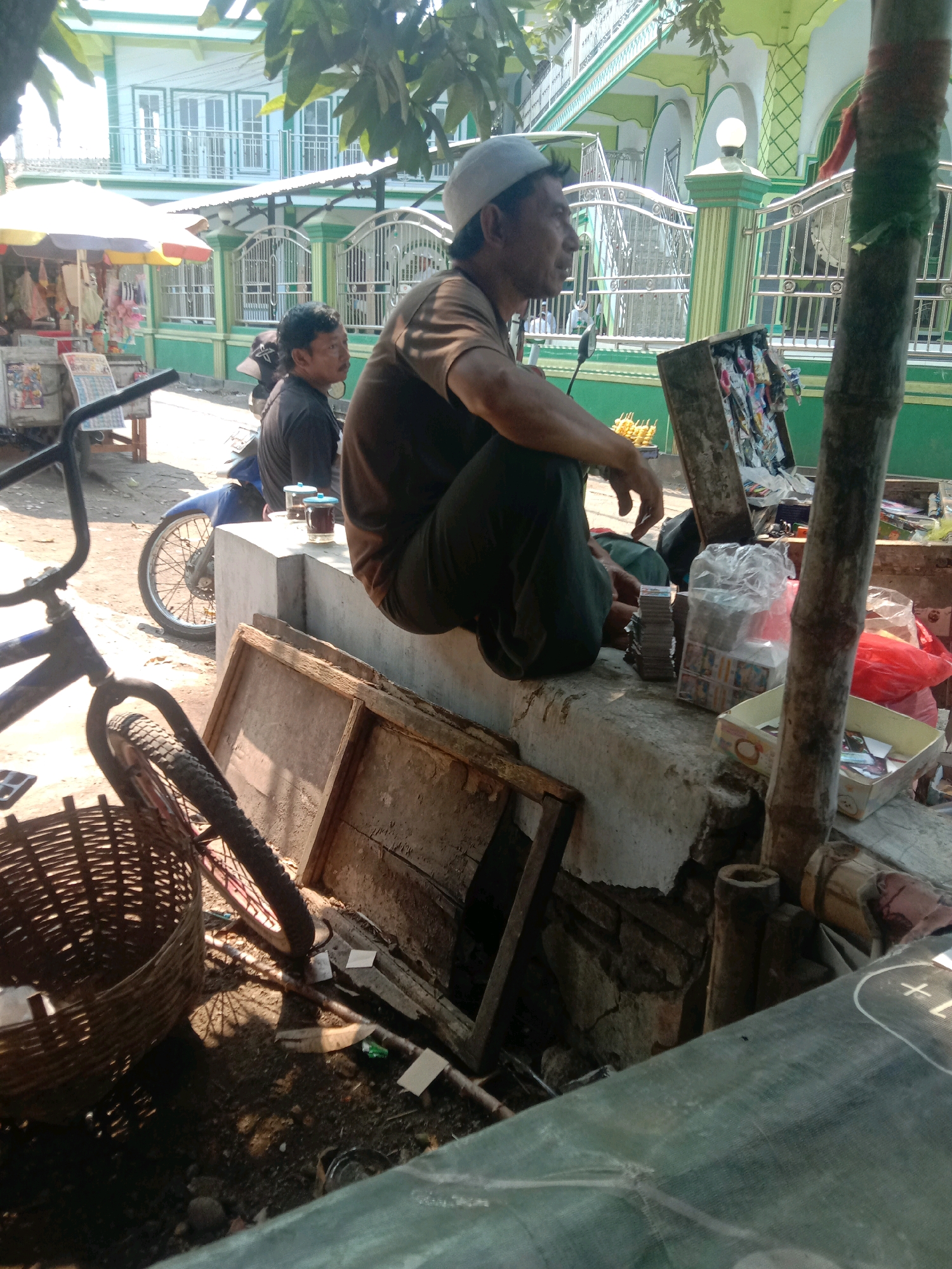 In the city of Ngempit, Indonesia, a man in a white cap and brown shirt sits on a concrete ledge outdoors, facing right, with his legs crossed. He appears to be looking at something out of frame to the right. Next to him, another man in a black shirt sits on a motorcycle. In front of the seated men, on the ledge, are two cups of dark liquid. To the left of the ledge, a bicycle with a basket is parked. Behind the ledge, a building with green and white accents and a white railing is visible, possibly a mosque. Various items are scattered around the ledge, including a wooden frame, a stack of cards, and plastic bags of goods. The ground is dirt and gravel, with some debris. The scene suggests a street-side vendor or a casual gathering in a public space.