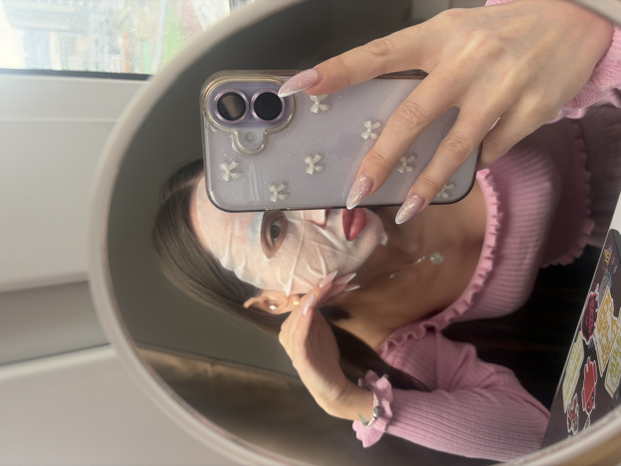 A woman wearing a pink ribbed top is taking a mirror selfie. Her face is covered with a white sheet mask. She holds an iPhone in her hand, which has a light purple case with small white flower embellishments. The phone's camera lenses are visible. Her nails are long, almond-shaped, and decorated with glitter polish. A silver necklace adorns her neck.
In the background, a window is visible, and the blurred outlines of a cityscape can be seen. Based on the style of the window and the architectural style visible, the location appears to be Saint Petersburg, Russia. The woman is likely inside a building, possibly her home or an apartment. A laptop covered in stickers is also visible.