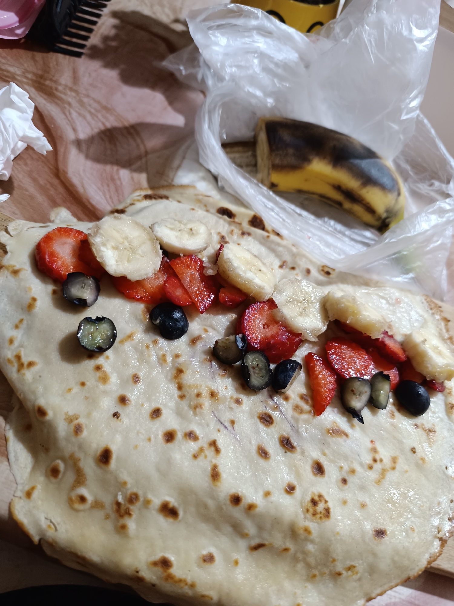 A crepe, garnished with strawberries, blueberries, and sliced bananas, sits on a wooden surface alongside a plastic bag containing a partially eaten, overripe banana. The scene also includes a hair comb and crumpled tissues. There's no explicit indication of it being in Pleven, Bulgaria, or any identifiable landmarks to verify this.
