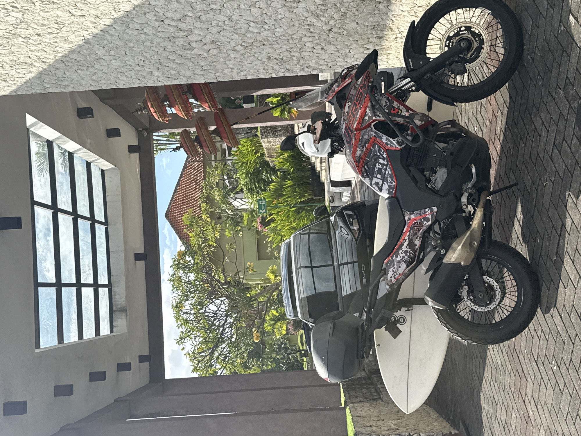In Kuta Selatan, Indonesia, a motorcycle with a white surfboard attached to its side is parked on a brick-patterned driveway. The motorcycle is red and black with a camouflage design, and a white helmet sits on its handlebars. Behind the motorcycle, a dark-colored SUV is parked. The scene is set in front of a building with a textured stone wall and a glass and metal roof structure. Lush green foliage and a terracotta-tiled roof are visible in the background. Several red and gold ornamental umbrellas hang from a pole near the building's entrance. The sun casts shadows across the pavement.