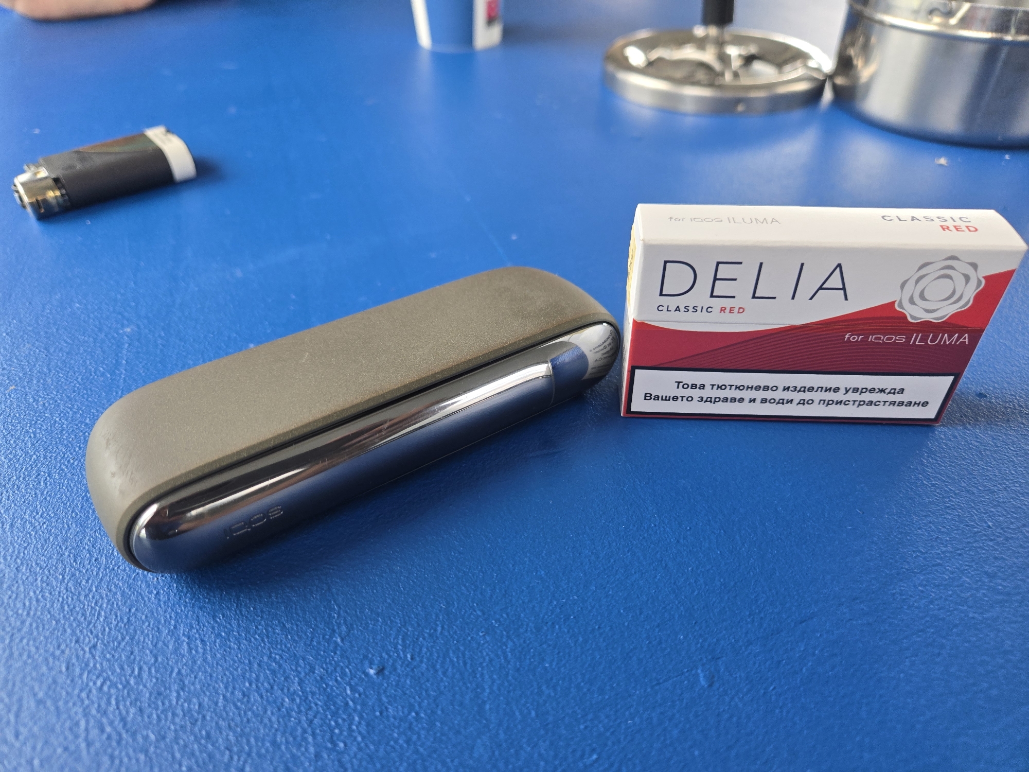 An overhead shot displays an IQOS ILUMA heated tobacco device and a pack of TEREA tobacco sticks on a vibrant blue, textured surface, likely a table.
In the center-left, the IQOS ILUMA device features a two-tone design: a matte grey charging case holding a sleek, metallic silver holder. The "IQOS" logo is subtly embossed on the metallic part of the holder. To its left, a discarded black and white cylindrical tobacco stick, consistent with a used TEREA stick, lies on the blue surface.
On the right side of the frame, a white and red pack of "DELIA CLASSIC RED" TEREA tobacco sticks for IQOS ILUMA is prominently displayed. The packaging clearly states "for IQOS ILUMA" multiple times, along with the product name "DELIA CLASSIC RED." A warning label in Bulgarian reads: "Това тютюнево изделие уврежда Вашето здраве и води до пристрастяване" (This tobacco product harms your health and leads to addiction).
In the upper right background, a metallic ashtray with a dark center is partially visible, alongside a glimpse of what might be a white cup. The lighting is bright and even, suggesting an indoor setting during the daytime. No people are visible, and no specific activity is captured, creating a static still life.