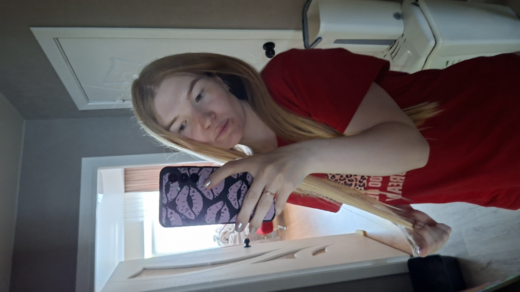 A woman is taking a selfie in a bathroom. The woman has long, strawberry blonde hair and is wearing a red t-shirt with black lettering. She is holding a smartphone with a purple case decorated with kiss prints. The bathroom has gray walls and white trim. In the background, there is a white door, a window with a partially-visible blind, a white cabinet above a toilet, and a wooden floor. The location is potentially Chaikovsky, Russia.