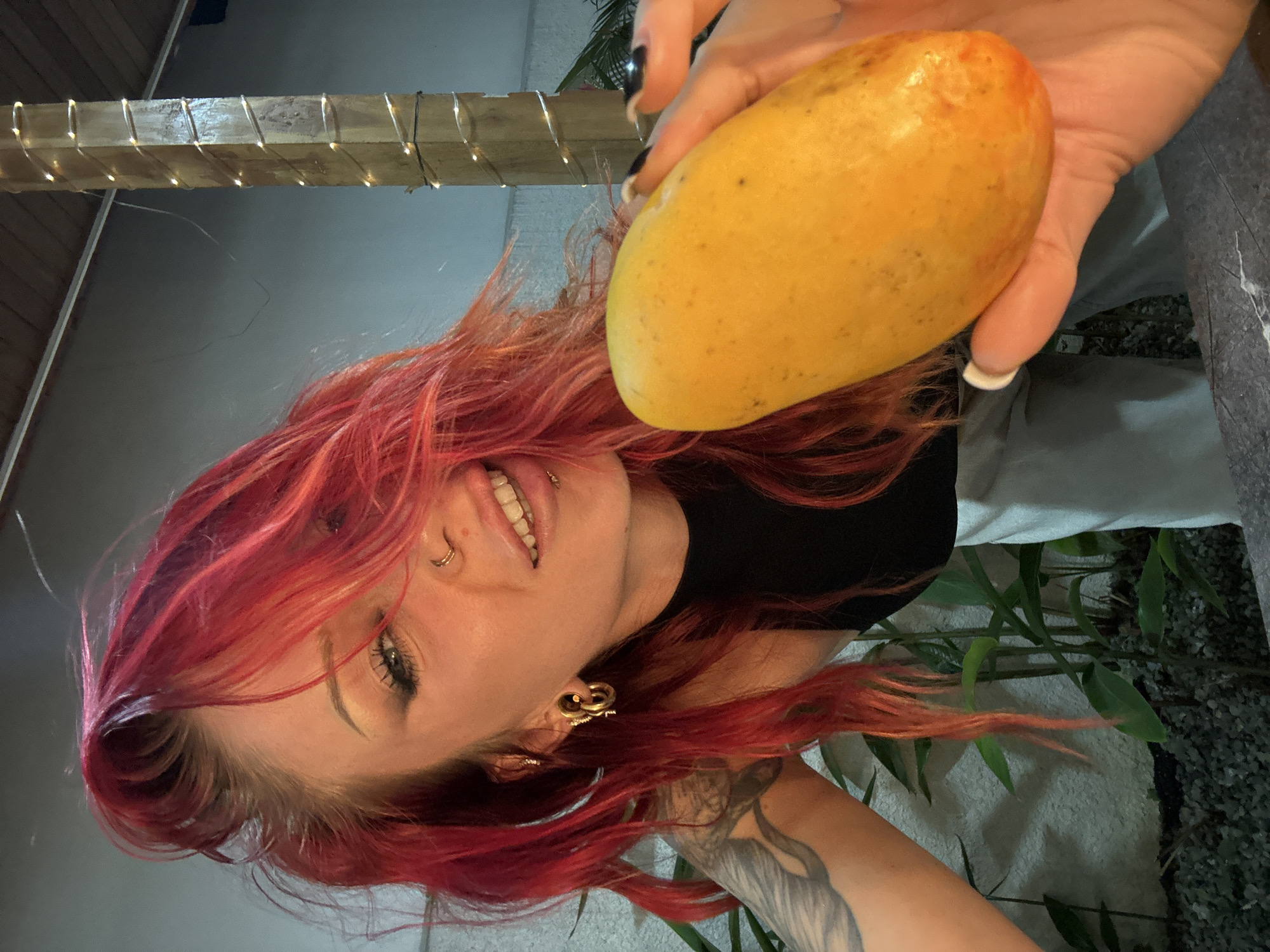 A young woman with vibrant red hair is holding a yellow mango up to the camera. She is wearing a black top and light-colored pants. Her hair is partially obscuring her face, but her left eye is visible, along with her nose piercing and a metal ring in her lip. She has large hoop earrings in her ears and a tattoo on her left arm. The background shows a wooden post wrapped with fairy lights, some lush green plants, and a gravel-covered ground. The scene appears to be outdoors, possibly in a tropical setting like North Kuta, Indonesia, given the vegetation and ambient lighting.