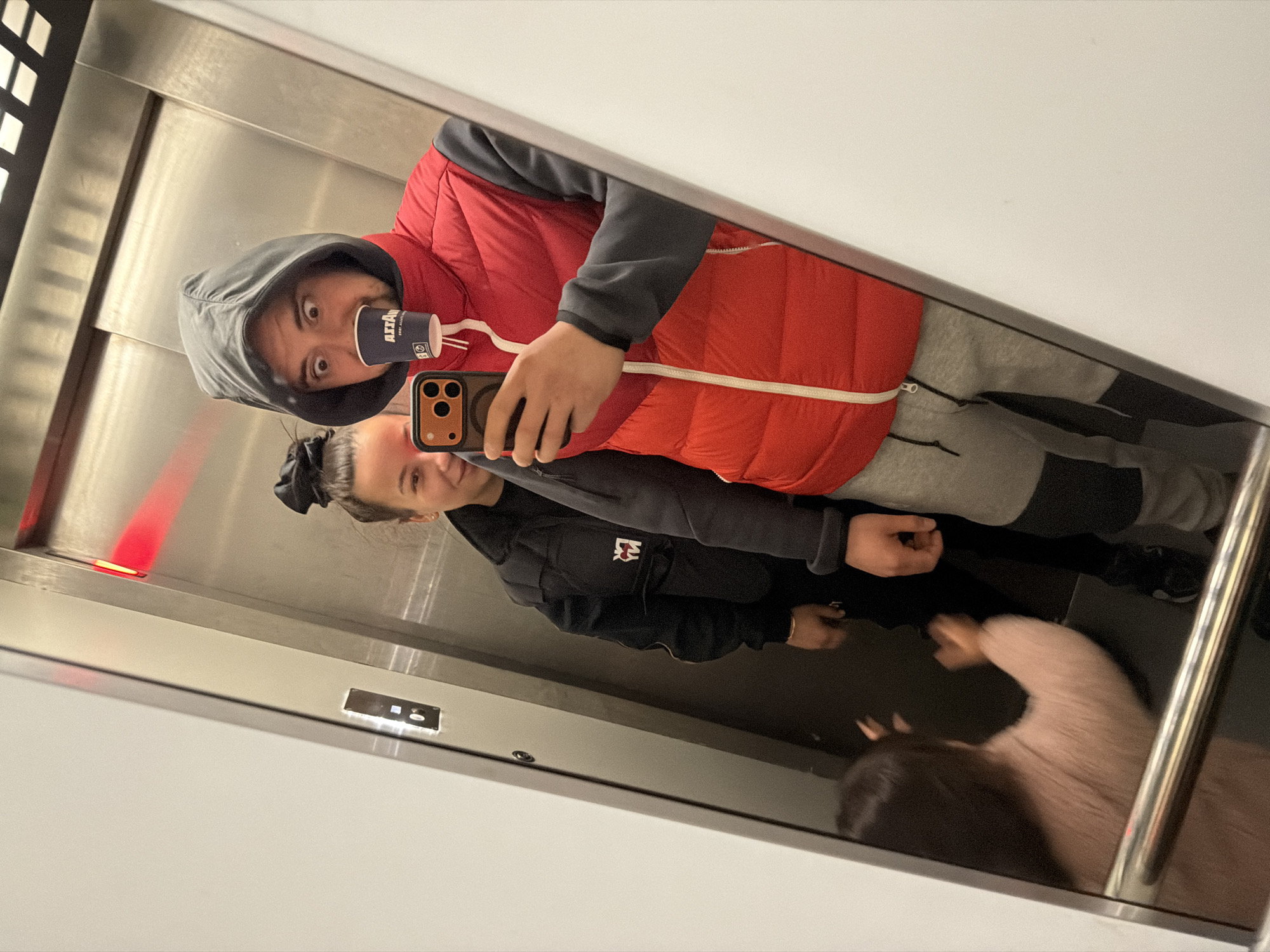 A selfie image shows a man, a woman, and part of a child in the reflection of a mirror in what appears to be an elevator. The man is wearing a hooded sweatshirt with a red vest over it, and holding a coffee cup and a cell phone. The woman is wearing a black jacket with a logo on the left side. The child has a light pink sweater. The elevator has silver walls. The city is listed as Unknown, Unknown.