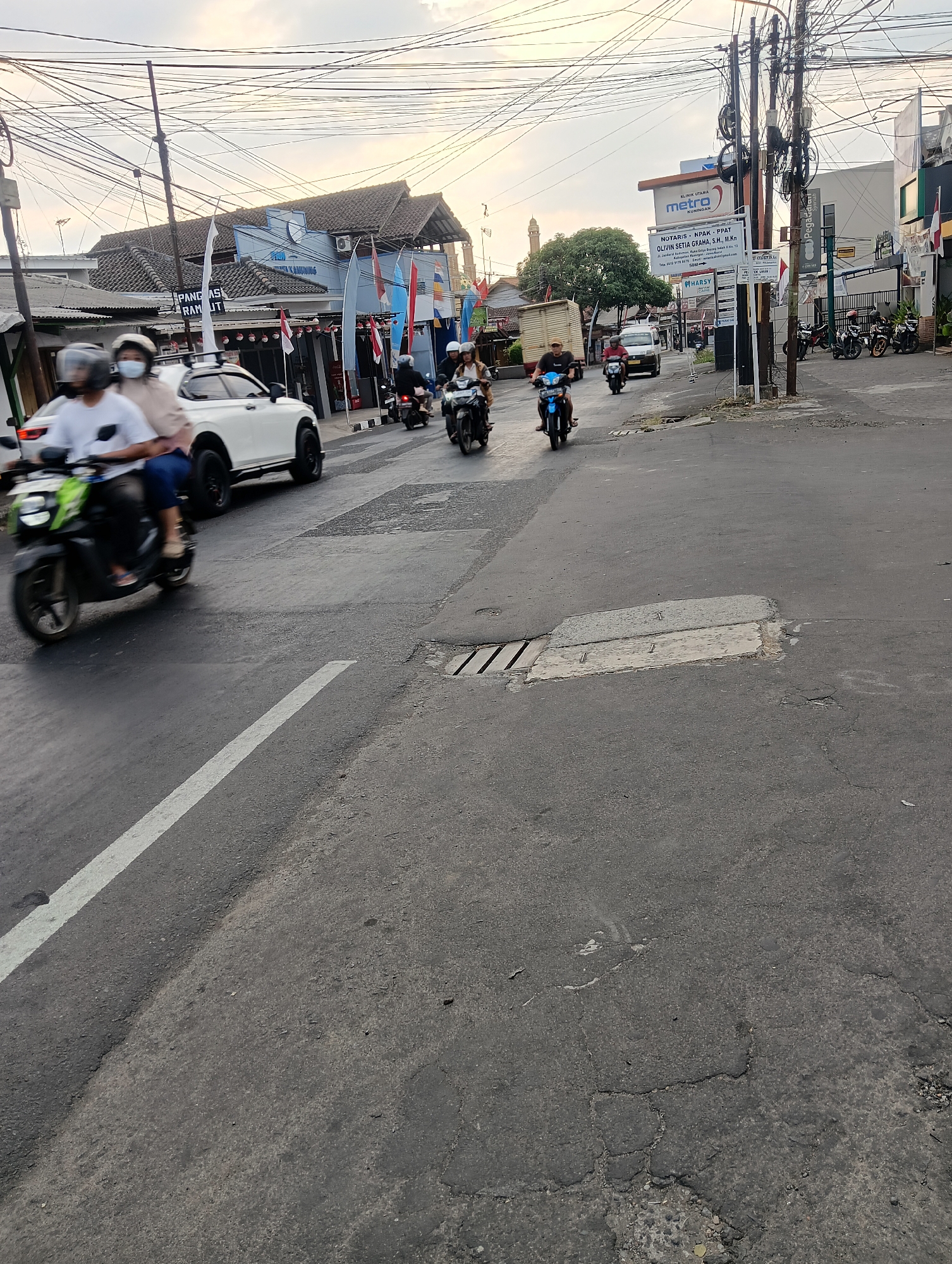 A street scene in Kuningan, Indonesia, features a two-lane asphalt road with a white painted lane divider. On the left side, a white SUV is parked while a motorcycle with two riders, both wearing helmets, passes by. The rider in front is wearing a white shirt and dark pants, while the passenger behind wears a light-colored jacket and dark pants. The motorcycle itself is black with green accents. Further down the road, several other motorcycles with riders are visible, some in motion. The background on the left shows a blue building with a tiled roof, adorned with flags and signs. On the right side of the street, a tall utility pole with numerous tangled wires stands prominently. Adjacent to it are buildings, including a modern building with a sign for "Pegadaian Syariah" and other commercial establishments, some displaying flags. A signpost with multiple advertisements and informational notices is also present. The sky above is overcast, suggesting either early morning or late afternoon. The overall scene depicts a typical urban street with active traffic and commercial activity.