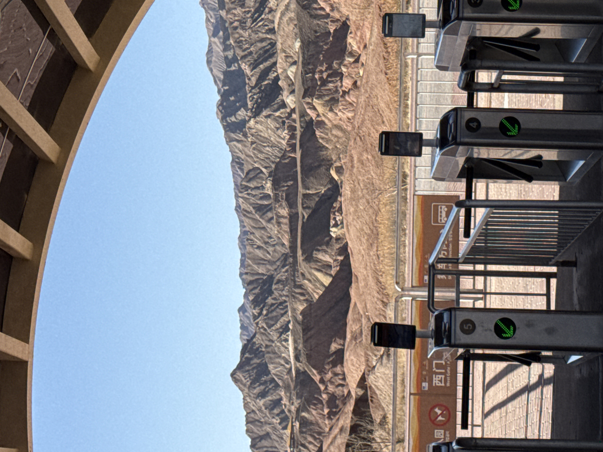 The image depicts a grand natural landscape viewed through an architectural structure, with a series of entry turnstiles in the foreground. The scene is captured on a clear, sunny day, suggested by the bright light and distinct shadows.
In the foreground, a set of modern, metallic turnstiles is visible. Each turnstile features a digital display showing a green circle with a downward-pointing arrow, indicating that passage is permitted. The turnstiles are numbered, with "3", "4", and "5" clearly visible. Behind the turnstiles, a light-colored, possibly brick-paved, ground stretches back. Several signs are placed near the gates. One sign clearly reads "West Gate A" with corresponding Chinese characters "西门 A" (Xīmén A). Another sign displays a bus icon, likely indicating transportation information.
The midground is dominated by a vast and rugged mountain range or canyon system, stretching horizontally across the frame. The mountains display arid, stratified rock formations in various shades of brown, tan, and reddish-brown, indicative of a dry, desert-like environment. The rock faces show deep crevices and natural erosion patterns.
Above the mountains, a clear, light blue sky fills the upper portion of the frame. To the far left, an architectural element, possibly part of an overhead walkway or an arched entrance, is partially visible. This structure is made of a light-colored material with visible beams and a curved design.
The overall scene suggests the entrance to a natural park or scenic area in Nantai, China, such as a major canyon or mountain tourist attraction, ready to welcome visitors. There are no people visible in the image.