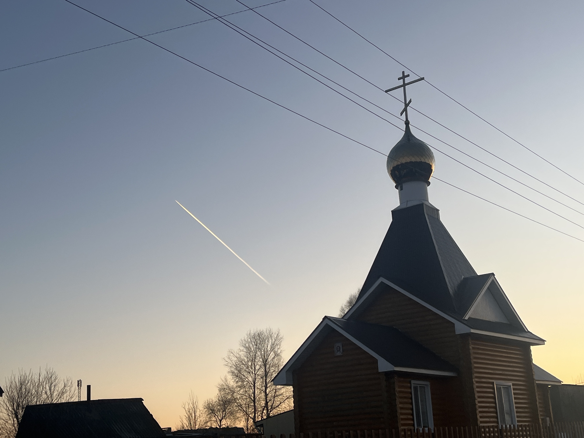 The image shows a wooden Orthodox church in Павловка, Russia, at sunrise or sunset. The church has a dark pyramidal roof topped with a gold dome and a cross. Power lines are visible crossing the upper portion of the frame. A contrail from an airplane is also present in the sky. In the foreground, the roofs of other buildings and bare trees can be seen.
