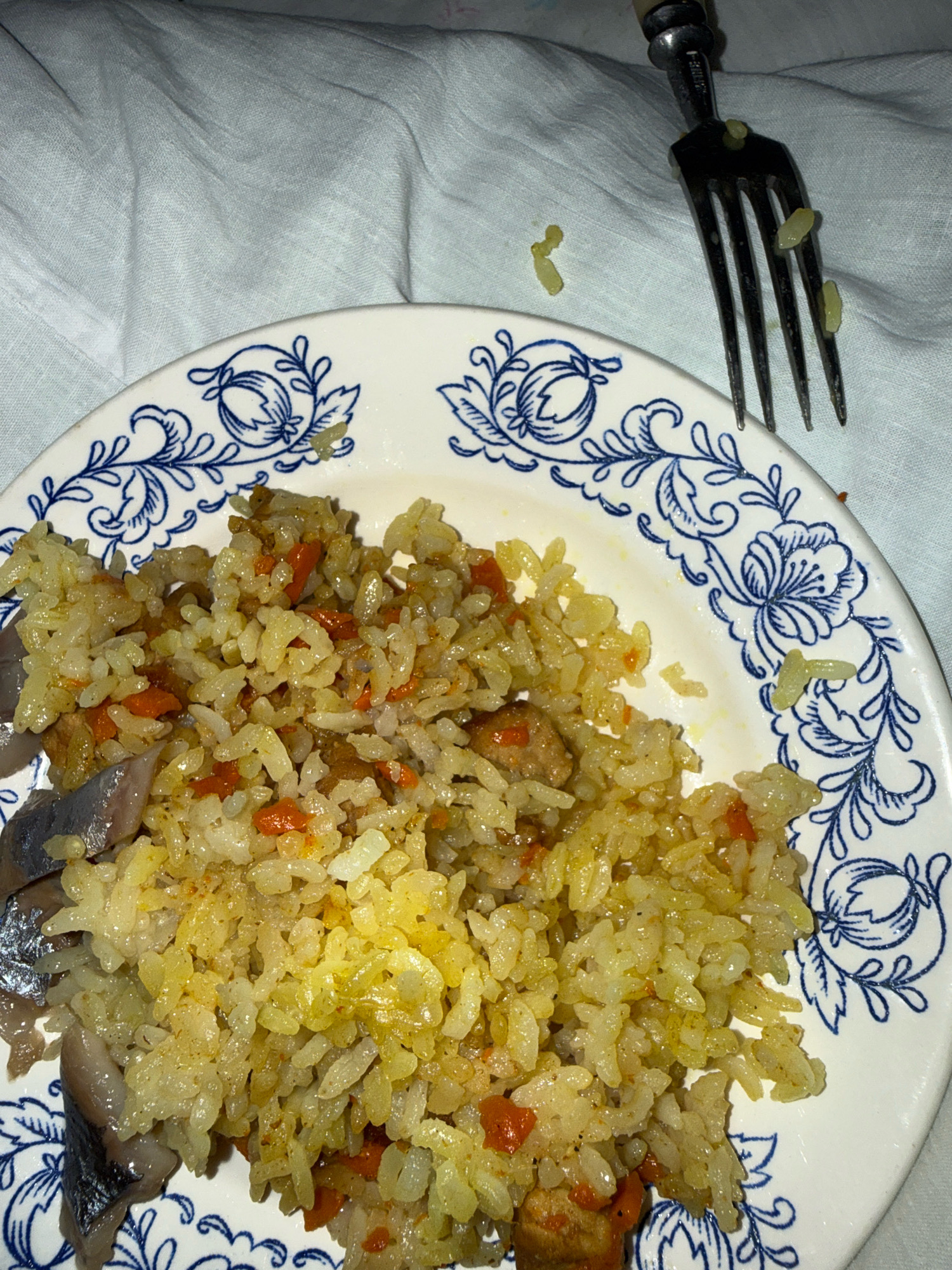 An overhead, medium shot captures a close-up of a plate of food, specifically pilaf with fish, placed on a wrinkled white cloth. A fork with a metallic handle and four tines is visible in the upper right corner, with a few pieces of rice on its tines and scattered around it. The plate is white with a decorative blue floral pattern around its rim. The pilaf itself is a mixture of rice, small pieces of orange carrot, and chunks of fish. The food appears to be on a table in a home setting, with the wrinkled white cloth suggesting it might be a tablecloth or bedsheet. No people are visible in the frame. The lighting is dim, creating some shadows and reflections on the food. The image was taken in the city of Zavolzhye, Russia.