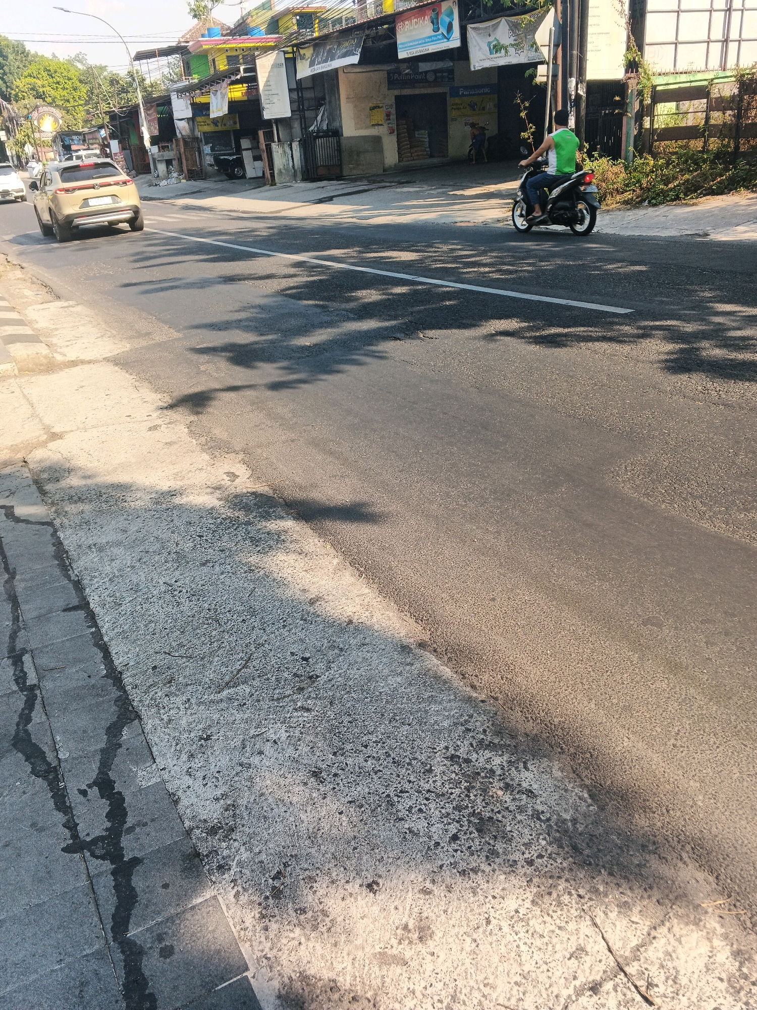 In Ungaran, Indonesia, a street scene unfolds with a beige SUV driving on the asphalt road. To the right, a person on a motorcycle wearing a green and white sleeveless shirt and blue jeans rides in the same direction as the SUV. The road is lined with sidewalks and buildings, with trees and vegetation visible in the background. The scene is bathed in sunlight, casting shadows on the road.
