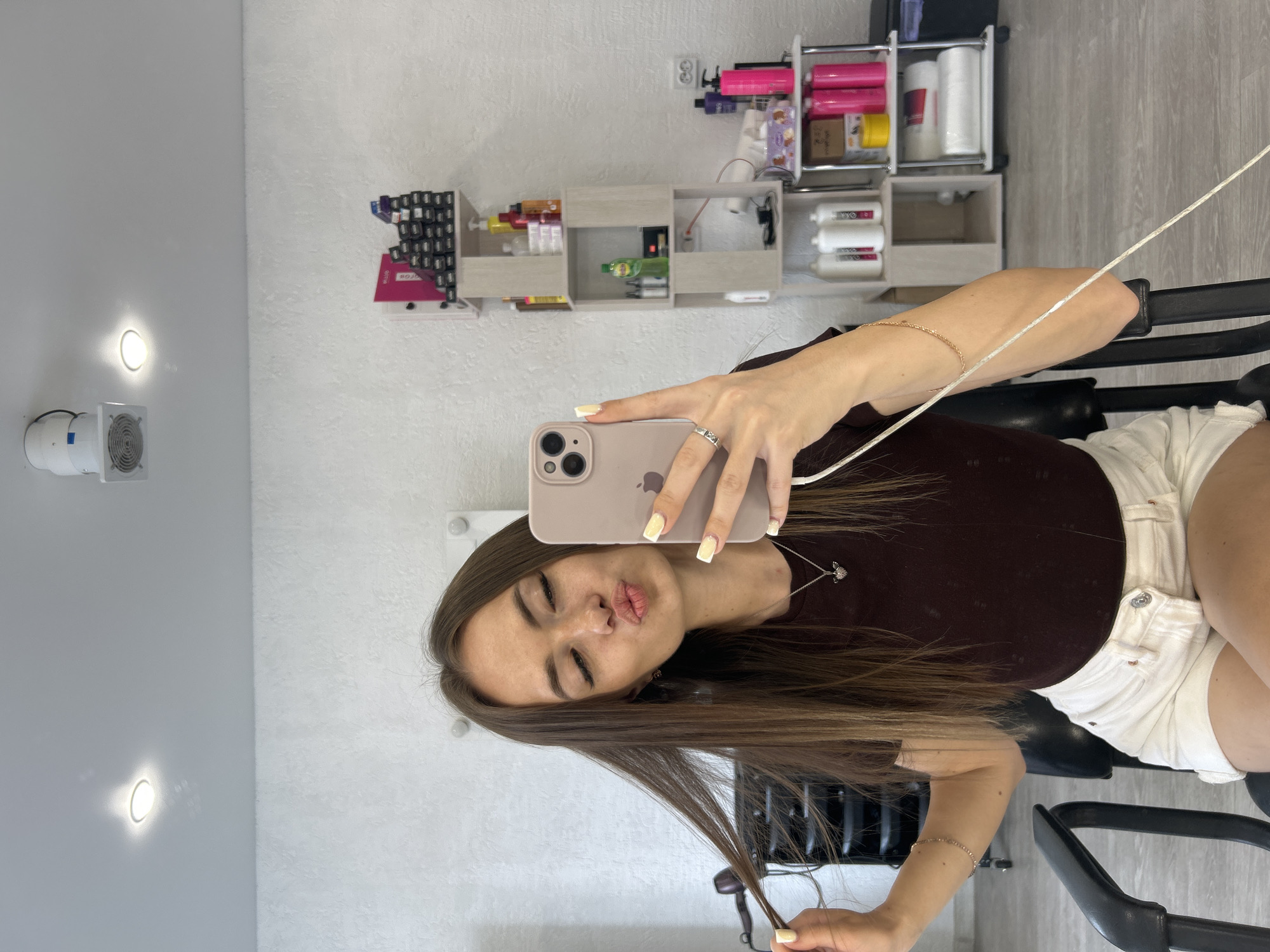 In a well-lit room, likely a salon given the shelving stocked with hair products, a young woman sits in a black chair. She is taking a selfie with a light pink smartphone, holding it with her right hand. She wears a dark maroon t-shirt and white shorts. Her long, brown hair is partially pulled to her left side. She has a silver necklace with a heart pendant and a silver ring on her right ring finger. Her fingernails are painted a pale yellow. The background shows white walls, a white ceiling with lighting fixtures, and shelves filled with various bottles and containers of beauty products.