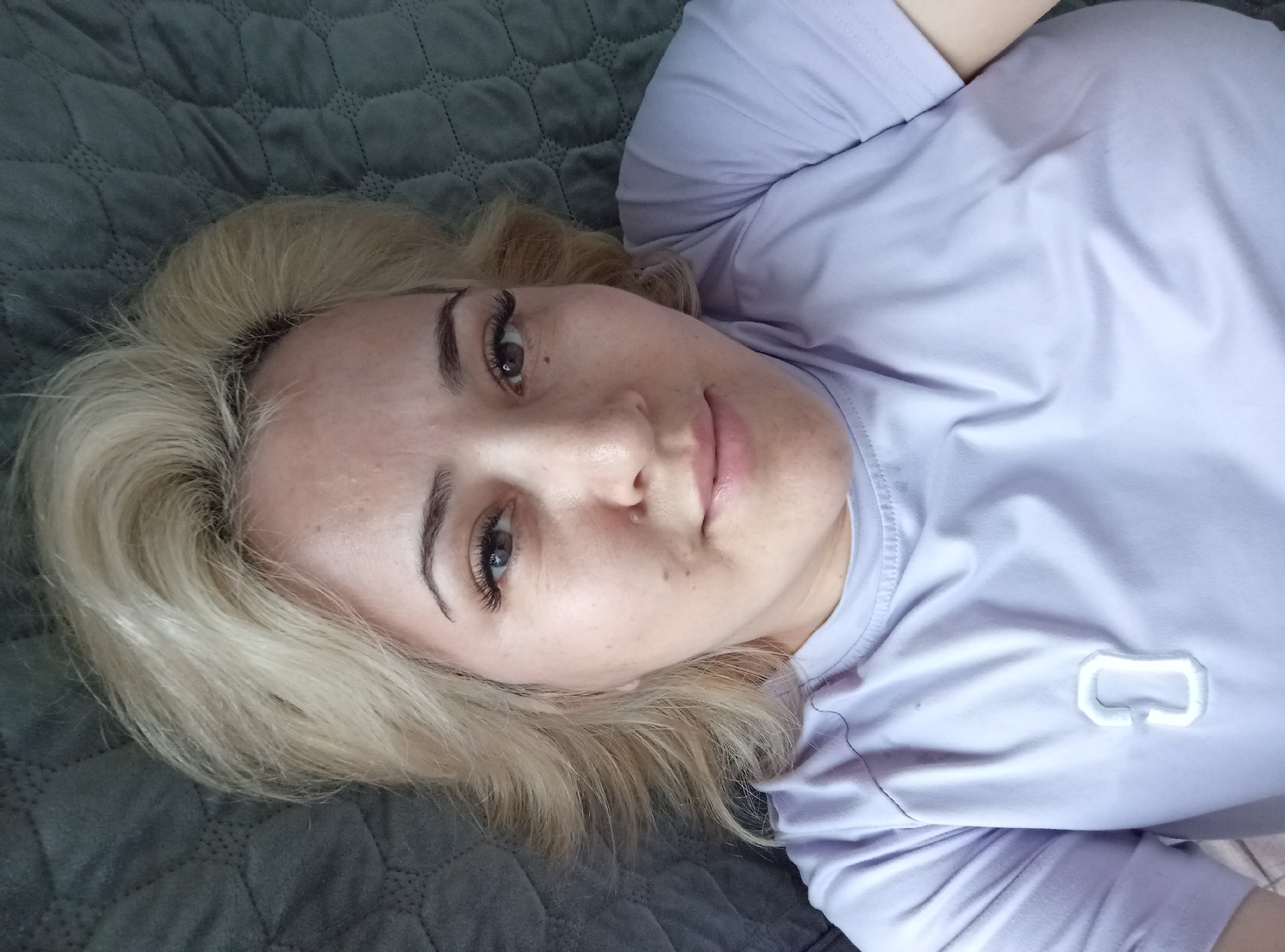 A woman with blonde hair and light skin lies on a dark gray quilted surface, possibly a bed or sofa. She is wearing a light purple t-shirt with a white embroidered logo on the chest. The image appears to be a selfie, with the woman looking towards the camera. She has noticeable eyelashes and some freckles on her face. The lighting is soft and diffused. The background is out of focus, but suggests an indoor environment. The scene is observed from a high-angle perspective, looking down at the woman.