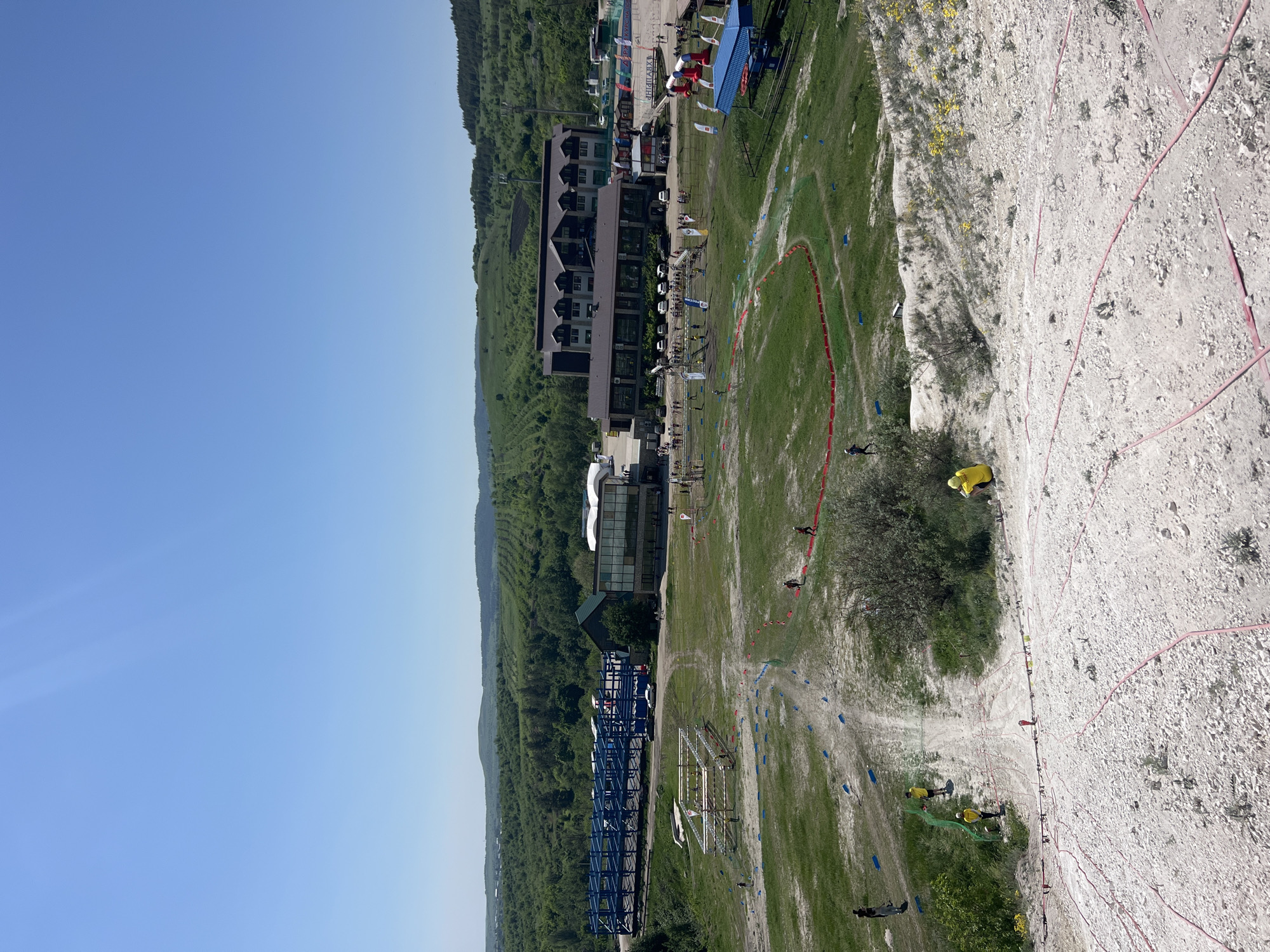 The image captures a scenic view from what appears to be a high vantage point in Khvalynsk, Russia. The landscape features a mix of natural terrain and man-made structures. A mountainous area covered with trees and green vegetation dominates the left side of the image, transitioning to a flatter, more open area in the center. Buildings, including a multi-story structure and some smaller structures, are situated in the middle ground, suggesting a developed or recreational area. The right side of the image presents a cliff face or steep slope, interspersed with vegetation and some climbing infrastructure like ropes. People are visible scattered throughout the scene, near the buildings, on the open ground, and near the cliff face. There is also a red border that makes a circle in the ground in the middle ground. The weather appears to be sunny, with clear skies overhead.