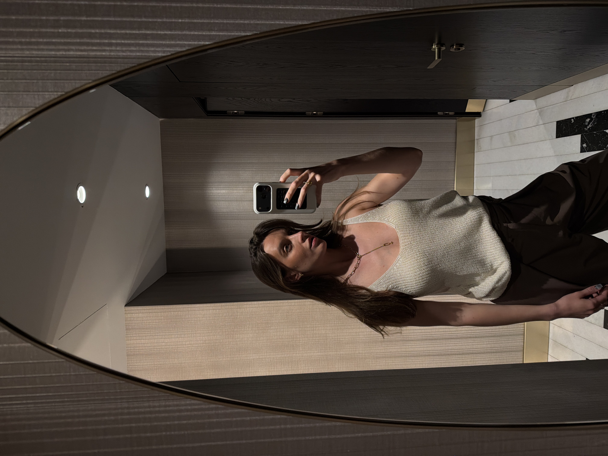 A woman is reflected in an oval mirror. She is taking a selfie, holding a white smartphone in her right hand. She is wearing a light-colored tank top and dark pants. The background shows a bathroom with light walls, a dark door, and marble tiles. No information indicates this is Budva, Montenegro.