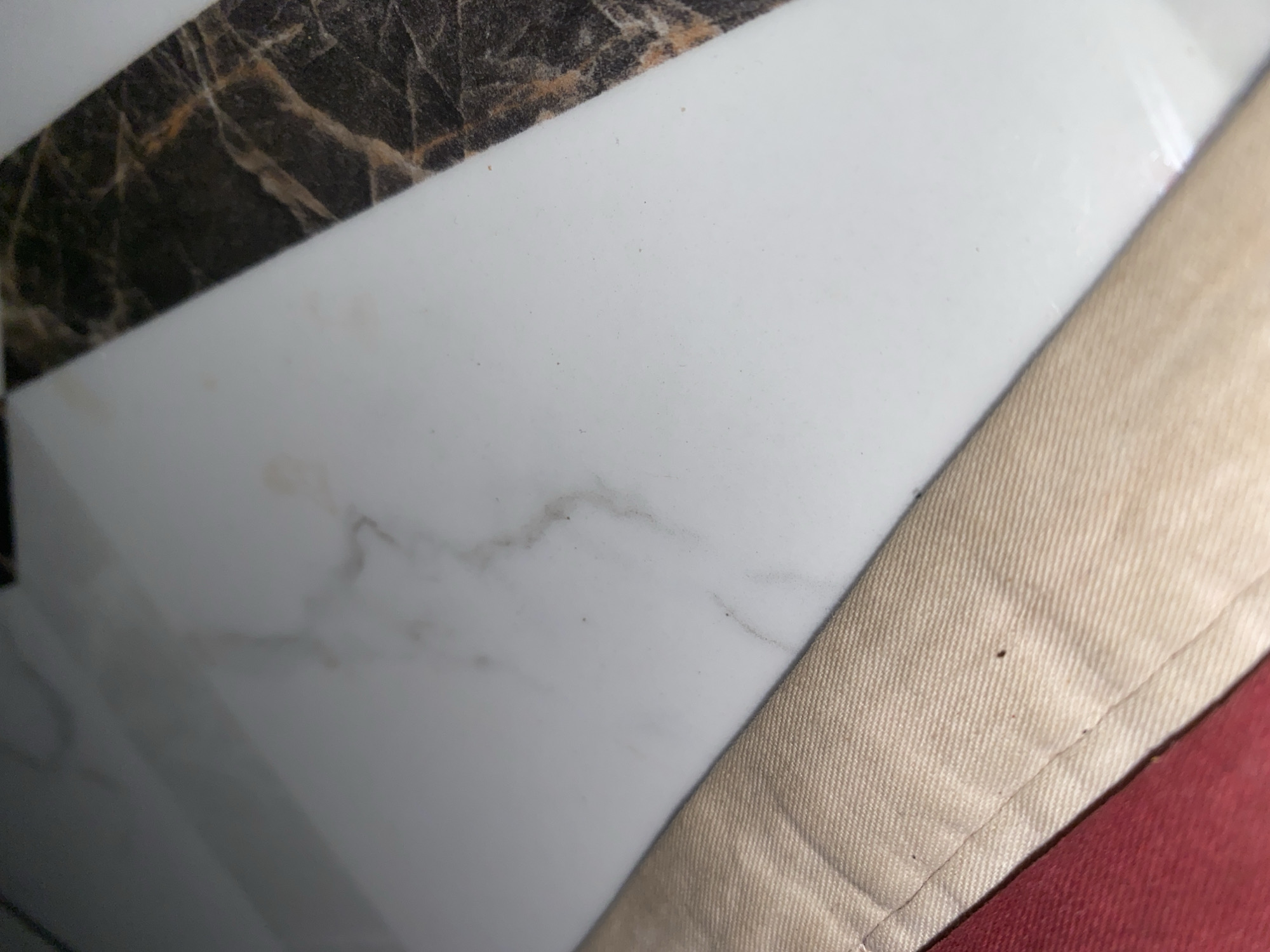 This is a close-up, overhead shot of a white, possibly marble surface with a dark, veined stone border. In the foreground, a beige fabric with visible stitching is angled across the frame, partially obscuring the white surface. A sliver of red fabric is visible at the bottom left edge of the image, beneath the beige fabric. The scene is domestic and does not depict any specific location in Jauro Yinu, Nigeria. There are no people or actions depicted.