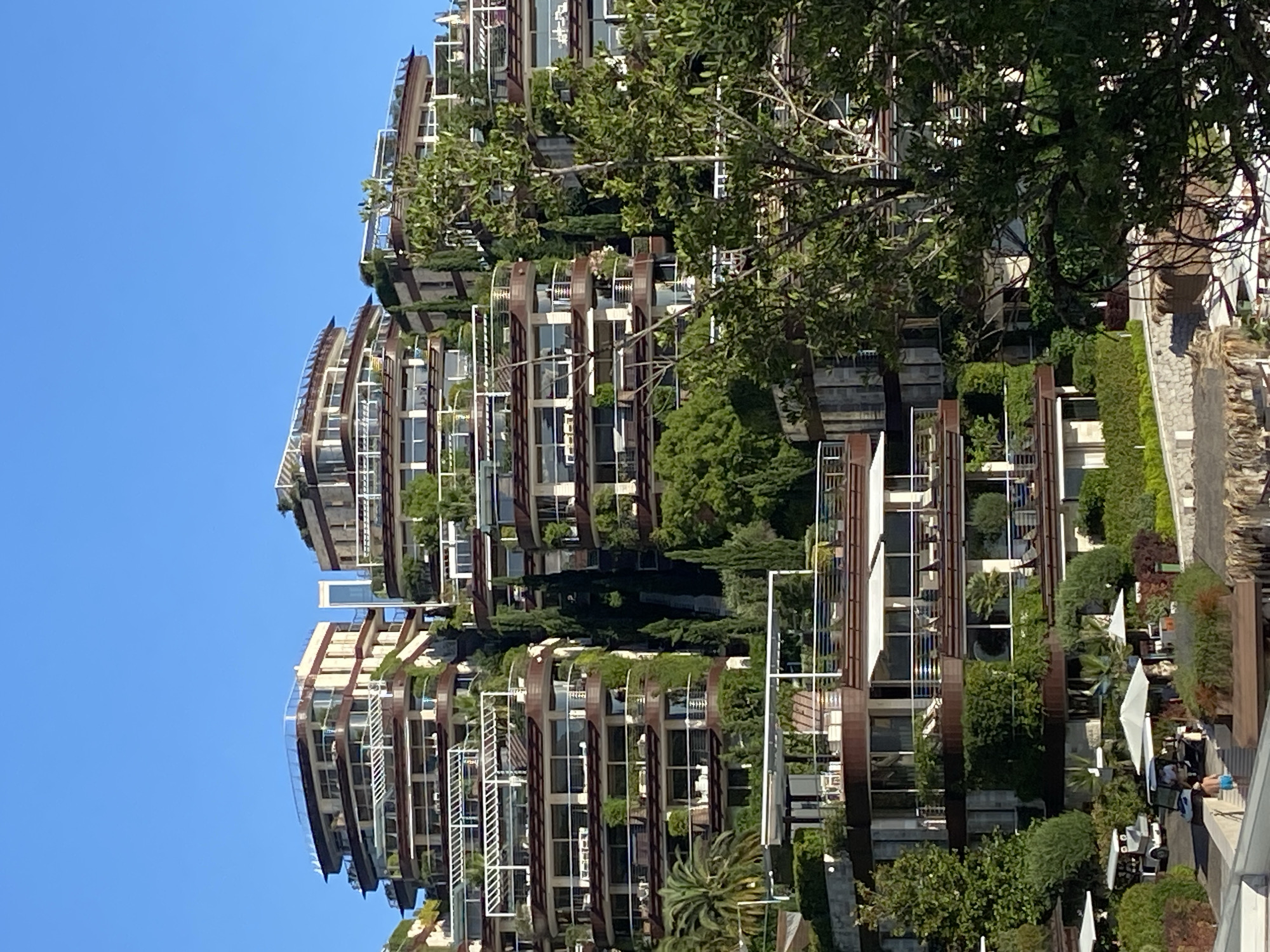 The image displays a terraced residential complex built into a hillside under a clear blue sky. The architecture features multiple levels with curved balconies, extensive glazing, and numerous green spaces, including trees, shrubs, and hanging plants integrated into the design. Several levels have awnings extended over balconies. In the foreground, a paved area with a few golf carts, umbrellas, and seating suggests an outdoor recreational or service area. Two individuals are visible near the golf carts. The overall impression is one of modern, luxurious living integrated with a natural landscape. This complex is likely located in Budva, Montenegro, known for its scenic coastal and hillside developments.