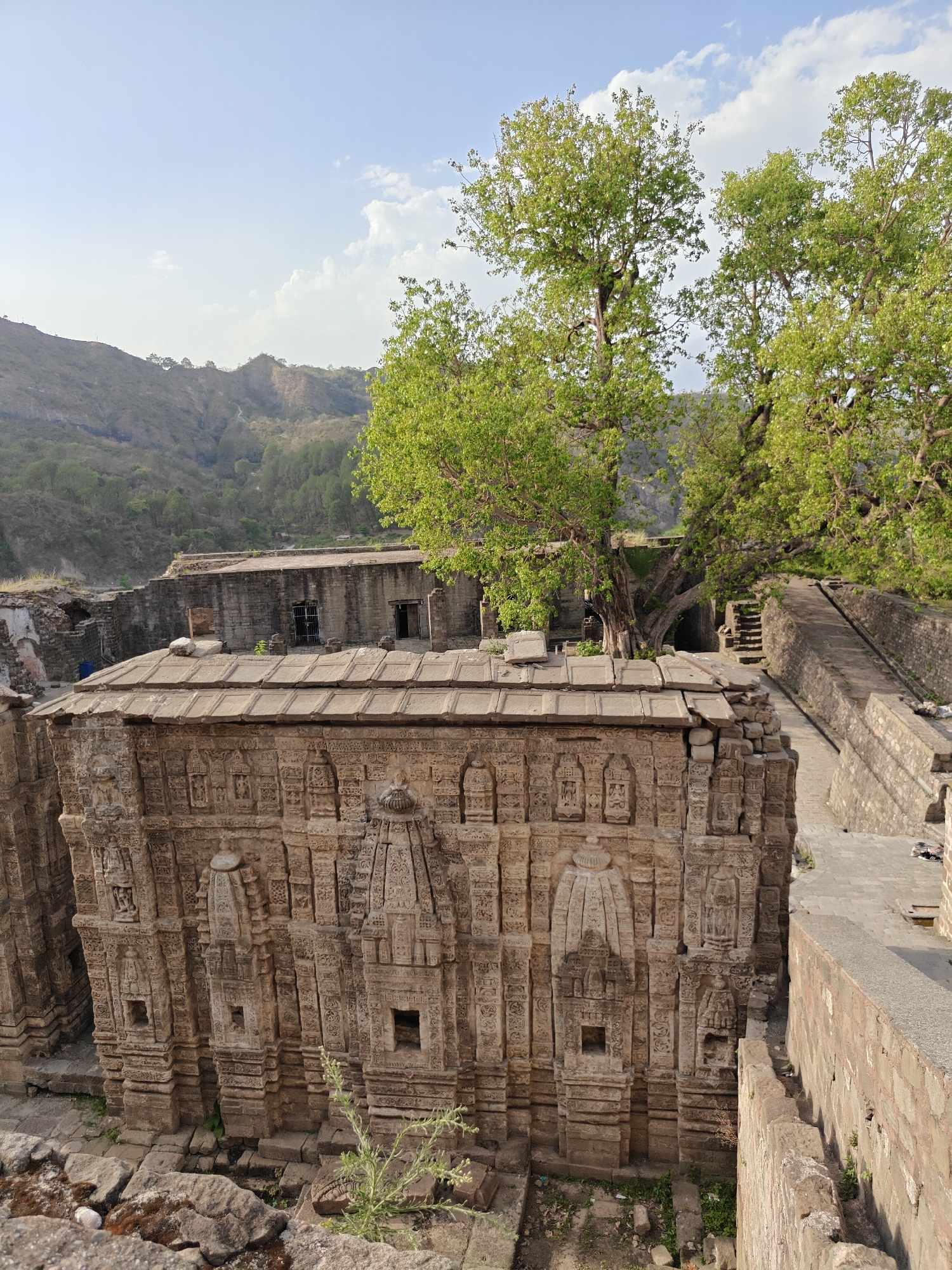 The image depicts the Masrur Temples, rock-cut Hindu temples located in the Kangra Valley of Himachal Pradesh, India, near the city of Kaswara. The primary focus is a large, elaborately carved stone temple structure, showing intricate details. The temple is built of sandstone. Several layers of carved details are observed on the outer wall, including miniaturized shrine-like projections. The temple's roof is made of stacked stone slabs.
Behind the temple, partially visible, is additional stone masonry, suggesting a larger complex. A pathway and staircase can be seen. Two large trees with green foliage are growing near the temple. The surrounding environment appears mountainous, with hills visible in the background. The sky is partly cloudy.