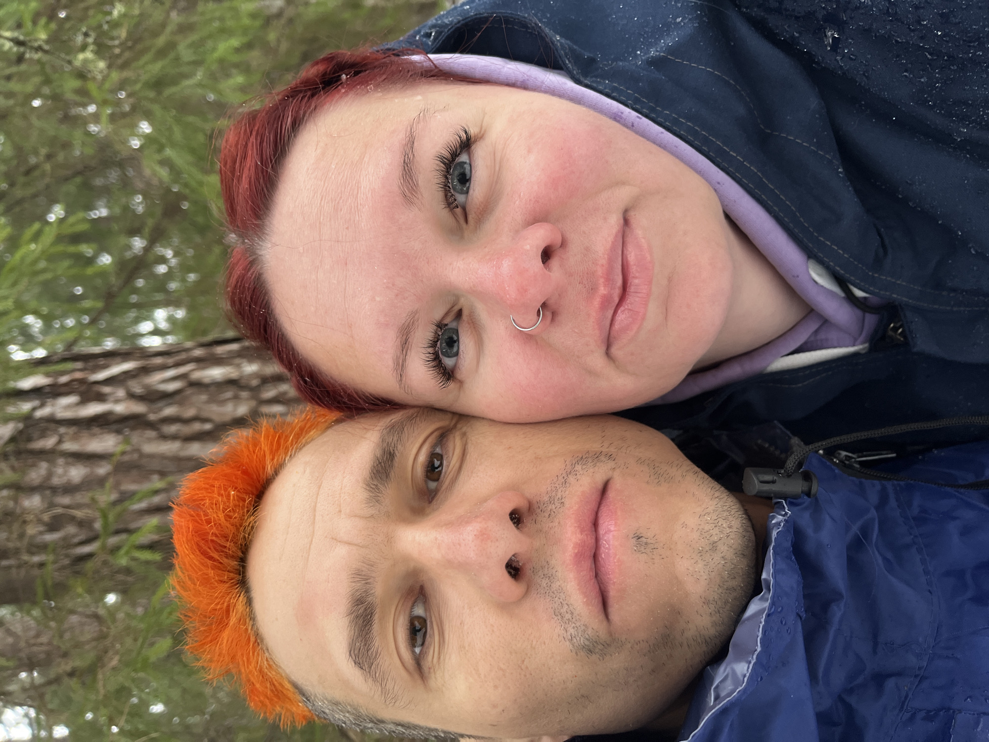 The image shows a close-up selfie of two people outdoors. The person on top has red hair, blue eyes, a nose ring, and is wearing a hooded jacket and a light purple hoodie. The person on the bottom has orange hair and brown eyes and is wearing a blue jacket. The background appears to be a tree. The location is Chaikovsky, Russia.