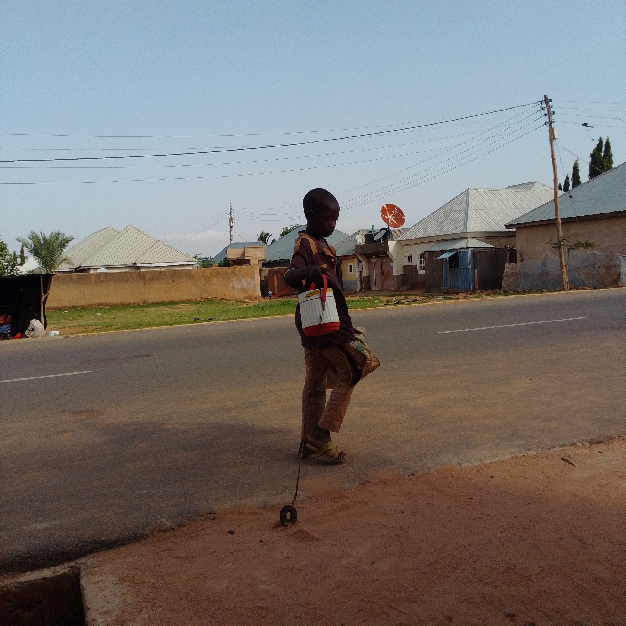 A young boy, appearing to be of African descent, walks on a dirt shoulder next to a paved road in Gombe, Nigeria. He is wearing a brown and yellow striped shirt and patterned brown trousers. In his right hand, he holds a red and white cylindrical container with a handle. A thin rope extends from the container down to a dark, circular object on the ground, appearing to be a toy or a training aid for rolling. Behind the boy, houses with silver corrugated metal roofs and a satellite dish are visible, situated behind a tan-colored wall. The sky is clear and light blue. The scene depicts a street in what appears to be a residential or mixed-use area of Gombe.