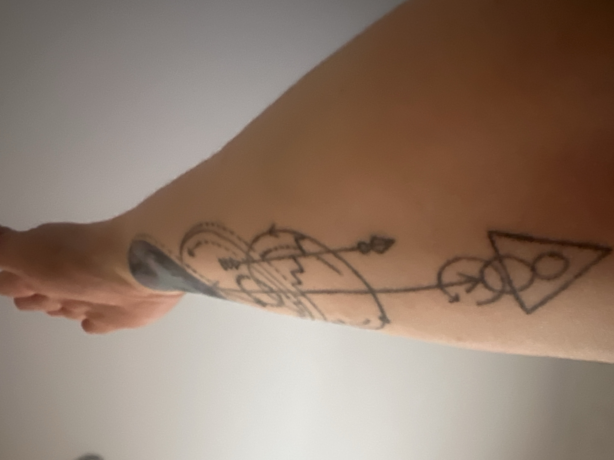 The image shows a person's arm with a tattoo. The tattoo appears to be a stylized design composed of geometric shapes, including circles, a triangle, and arrows, connected by lines. The shapes are rendered in black ink. No information in the image suggests the location is Chaikovsky, Russia.